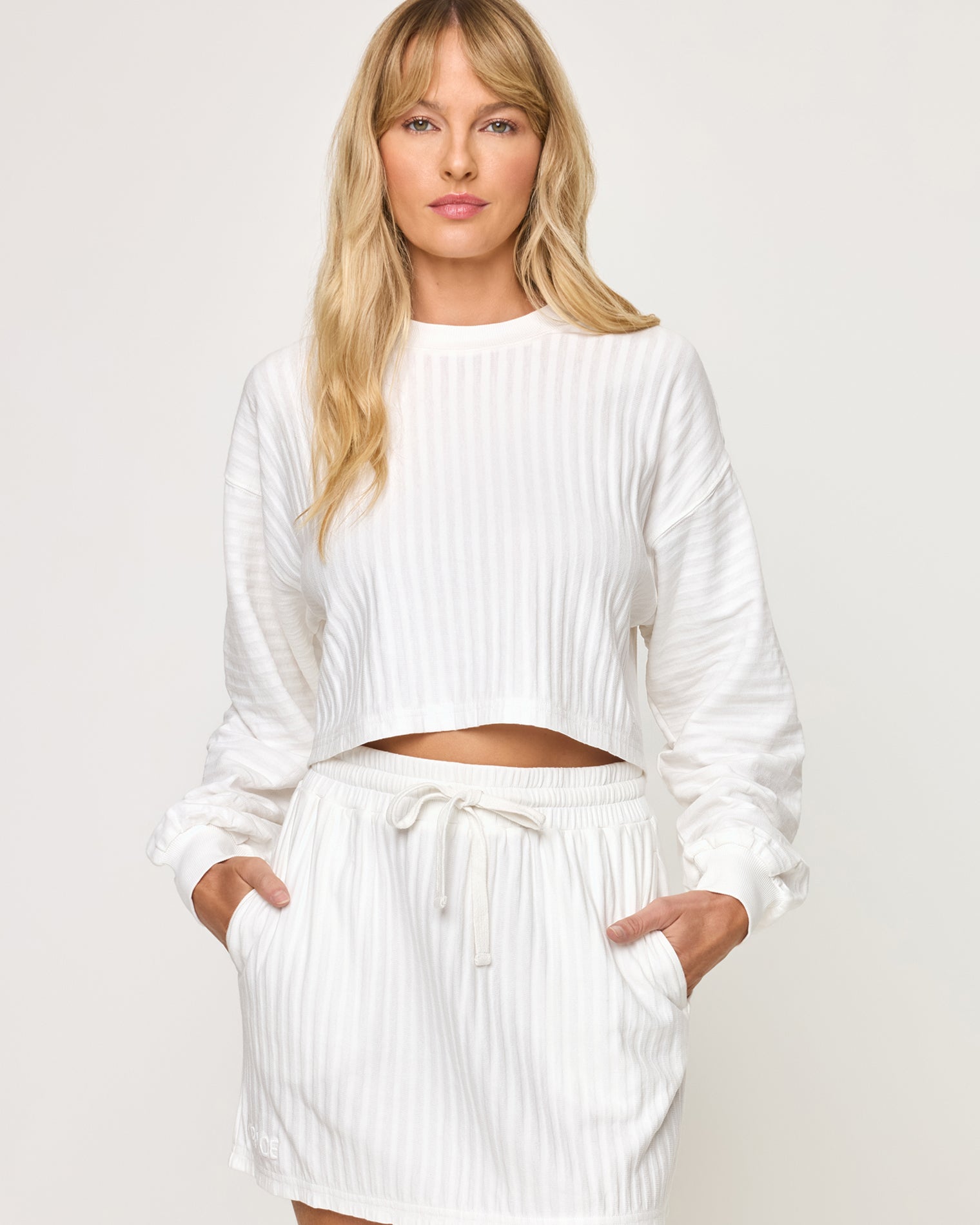Half Court Pullover - White Close-up front view of model wearing the Half Court Pullover in white, a ribbed long-sleeve cropped crewneck, with matching white Half Court Skirt, hands in pockets.