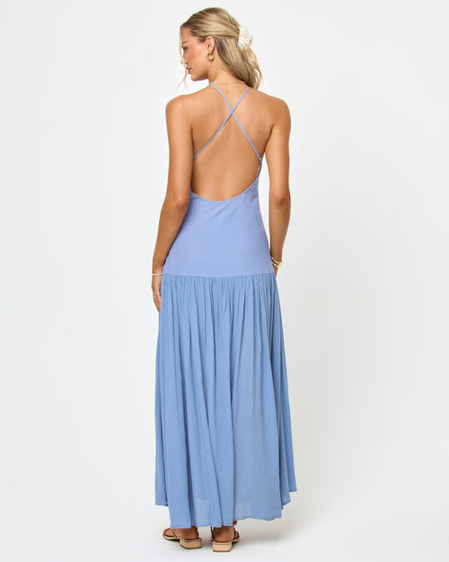 Back view of a model wearing the LSPACE Row Maxi Dress in chambray, showing the open back with crossback straps and a flowy pleated maxi skirt in light blue.