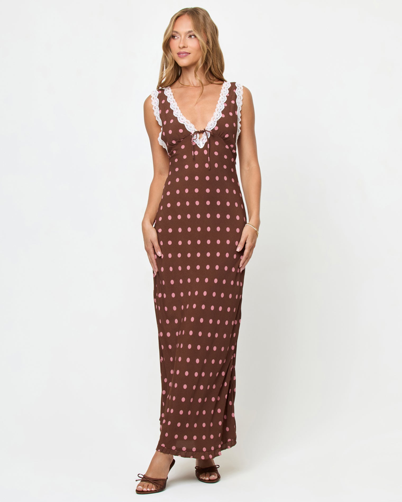 Eyes On You Maxi - Alotta Polka Dot Front view of a model wearing the LSPACE Eyes On You Maxi in Alotta Polka Dot on a white background, a brown maxi dress with large pink polka dots, white lace trim V-neckline, tie-front waist, and side slit, styled with brown heeled sandals.