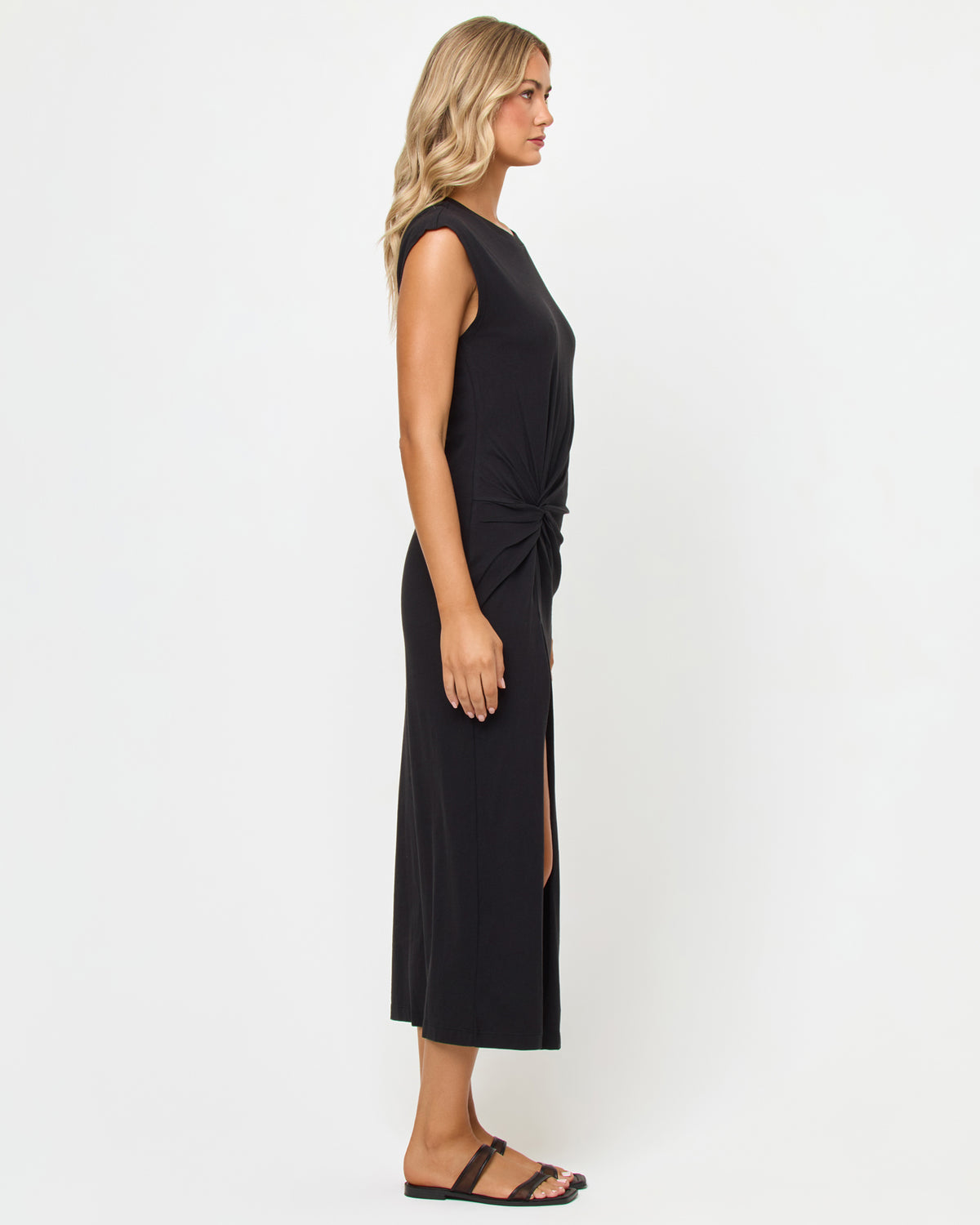 Sacha Maxi Dress - Black Side view of a model wearing the LSPACE Sacha Maxi Dress in black, showing the ruched twist detail at the hip, cap sleeves, and high side slit of the black sleeveless maxi dress, styled with black sandals.