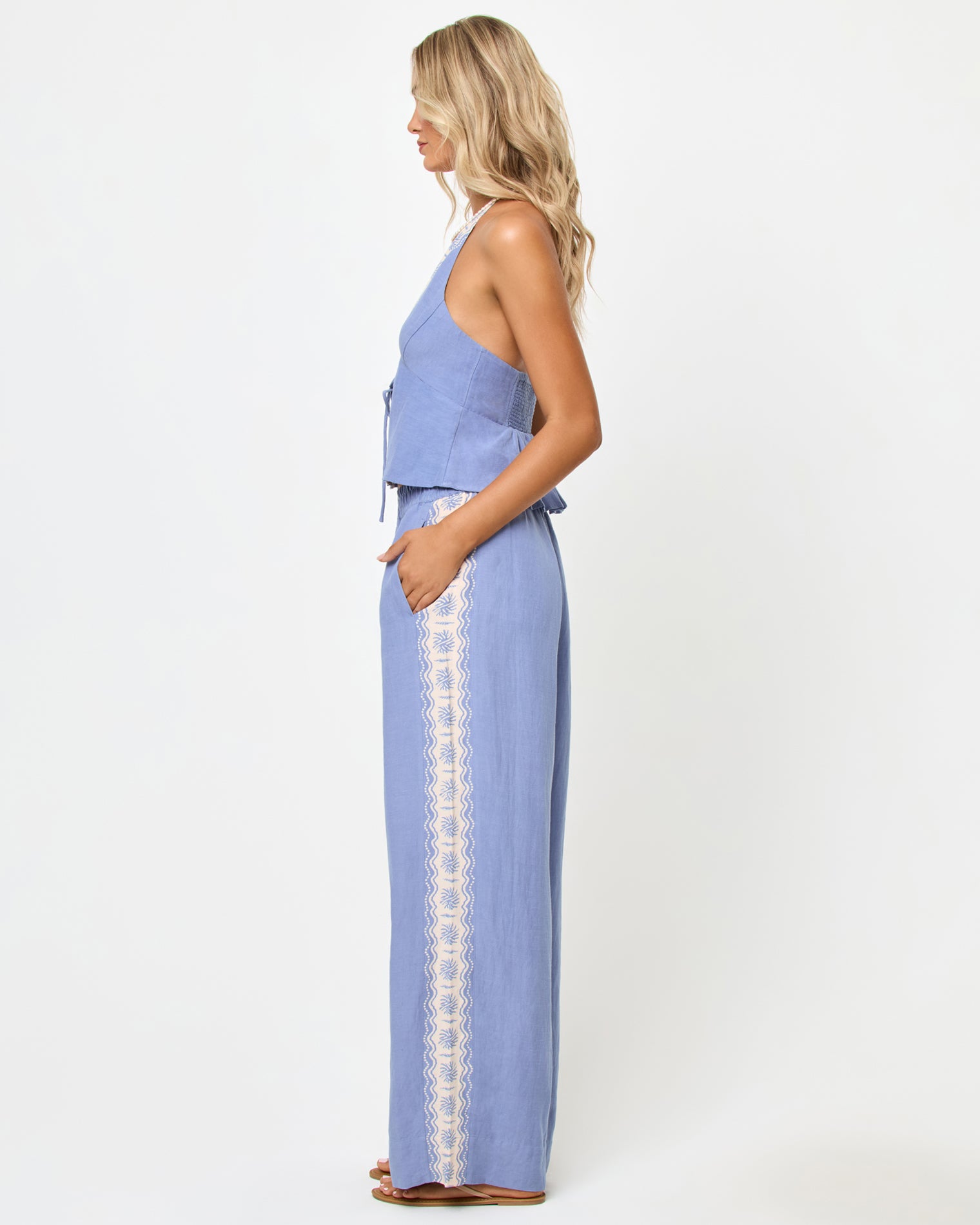 April Top - Shell Side view of a model wearing the LSPACE April Top in shell, a periwinkle blue halter top with a peplum ruffle hem, styled with matching wide-leg pants with cream embroidered trim.