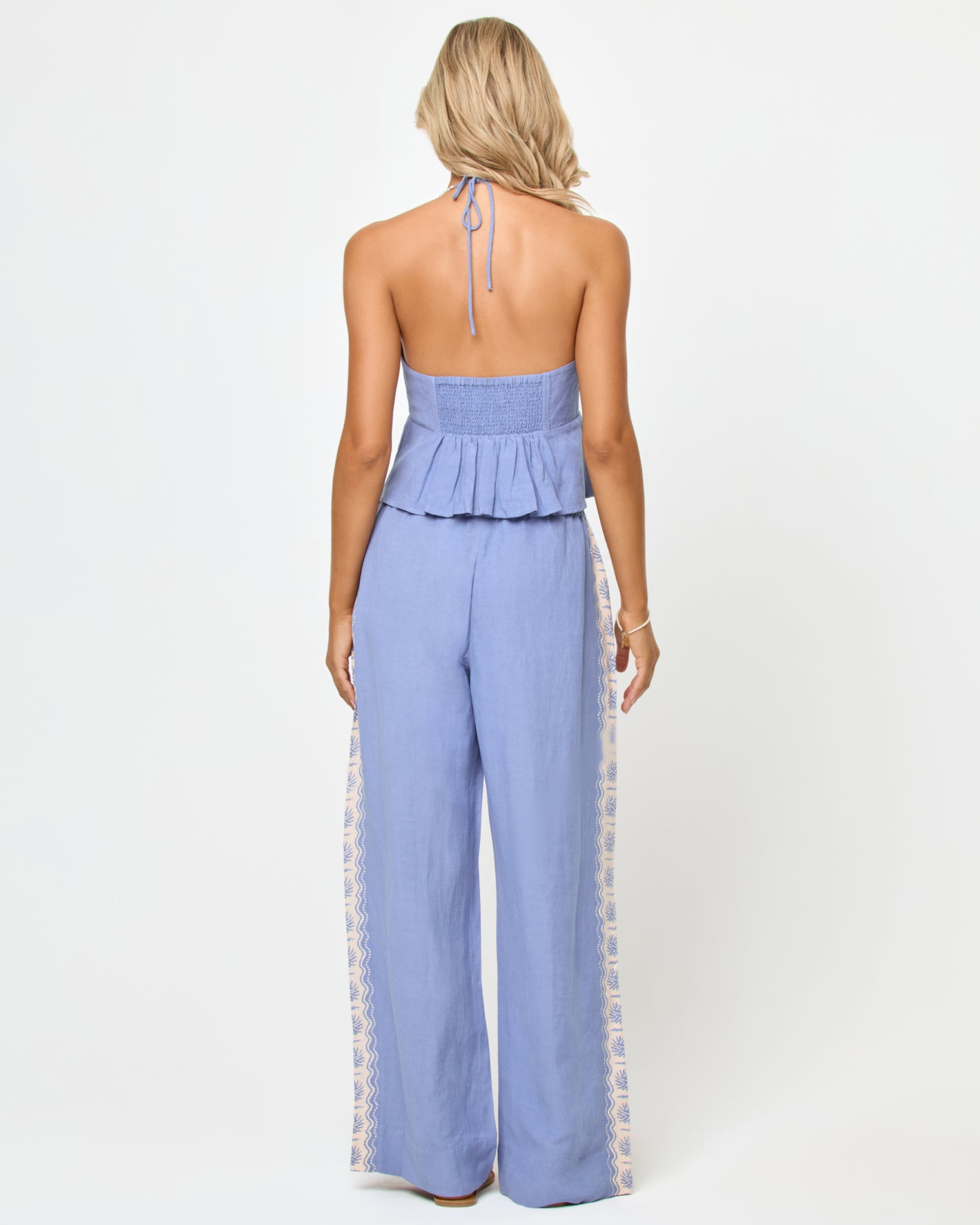 April Top - Shell Back view of a model wearing the LSPACE April Top in shell, showing the open back with halter neck tie, smocked band, and peplum ruffle hem, paired with matching wide-leg pants.