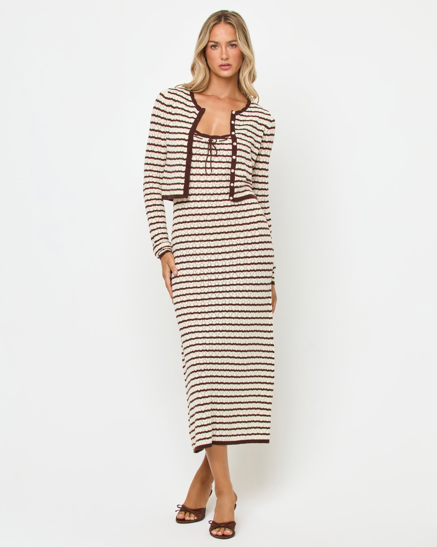 Rosalie Cardigan - Cafe-Cream Front view of a model wearing the LSPACE Rosalie Cardigan in cafe-cream, a cropped knit cardigan with cream and dark brown horizontal stripes and gold button closure, styled with the matching Rosalie Maxi dress and brown heels.