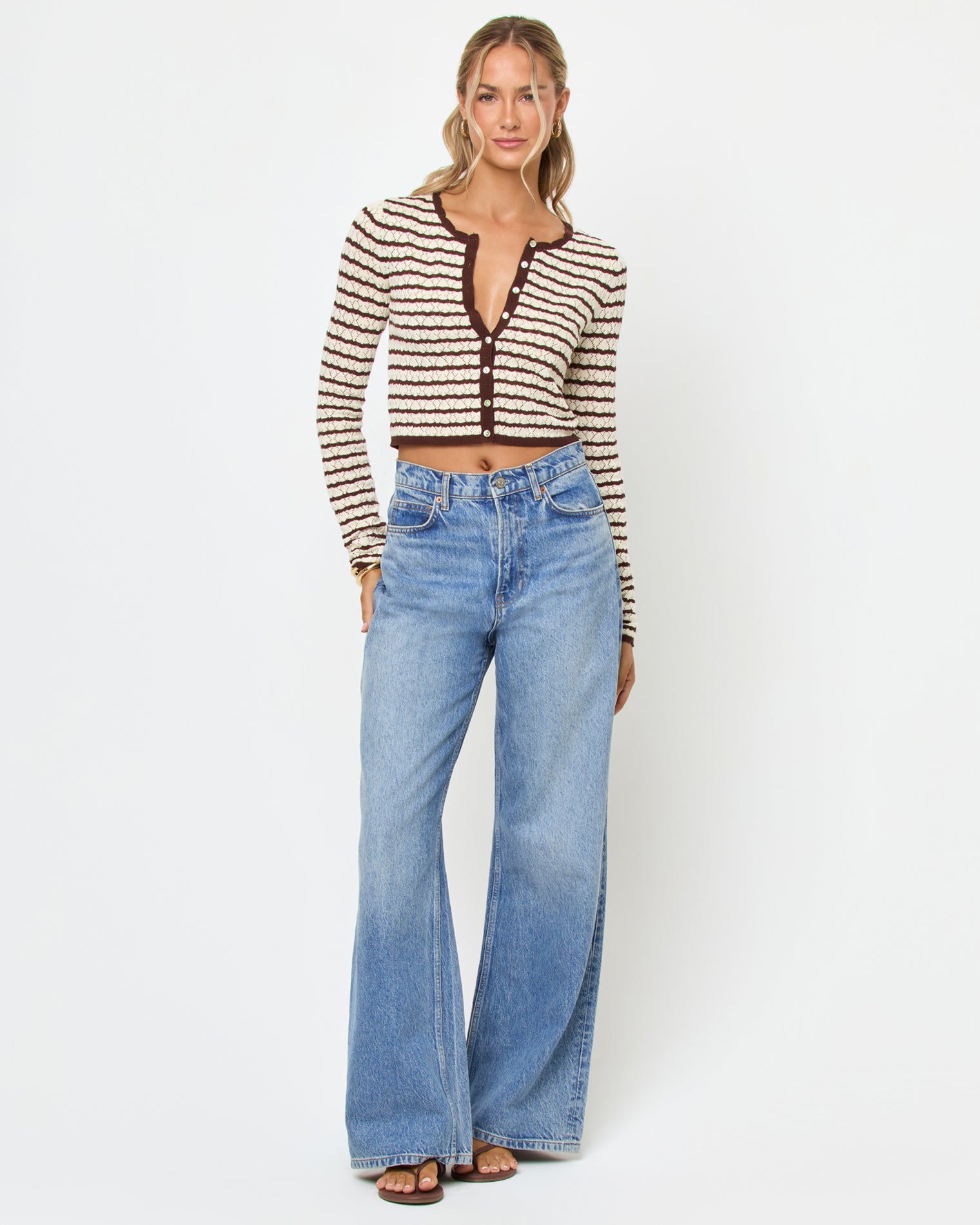 Rosalie Cardigan - Cafe-Cream Front view of a model wearing the LSPACE Rosalie Cardigan in cafe-cream, a cropped knit cardigan with cream and dark brown horizontal stripes, a V-neckline, and gold button closure, styled with wide-leg light wash jeans.