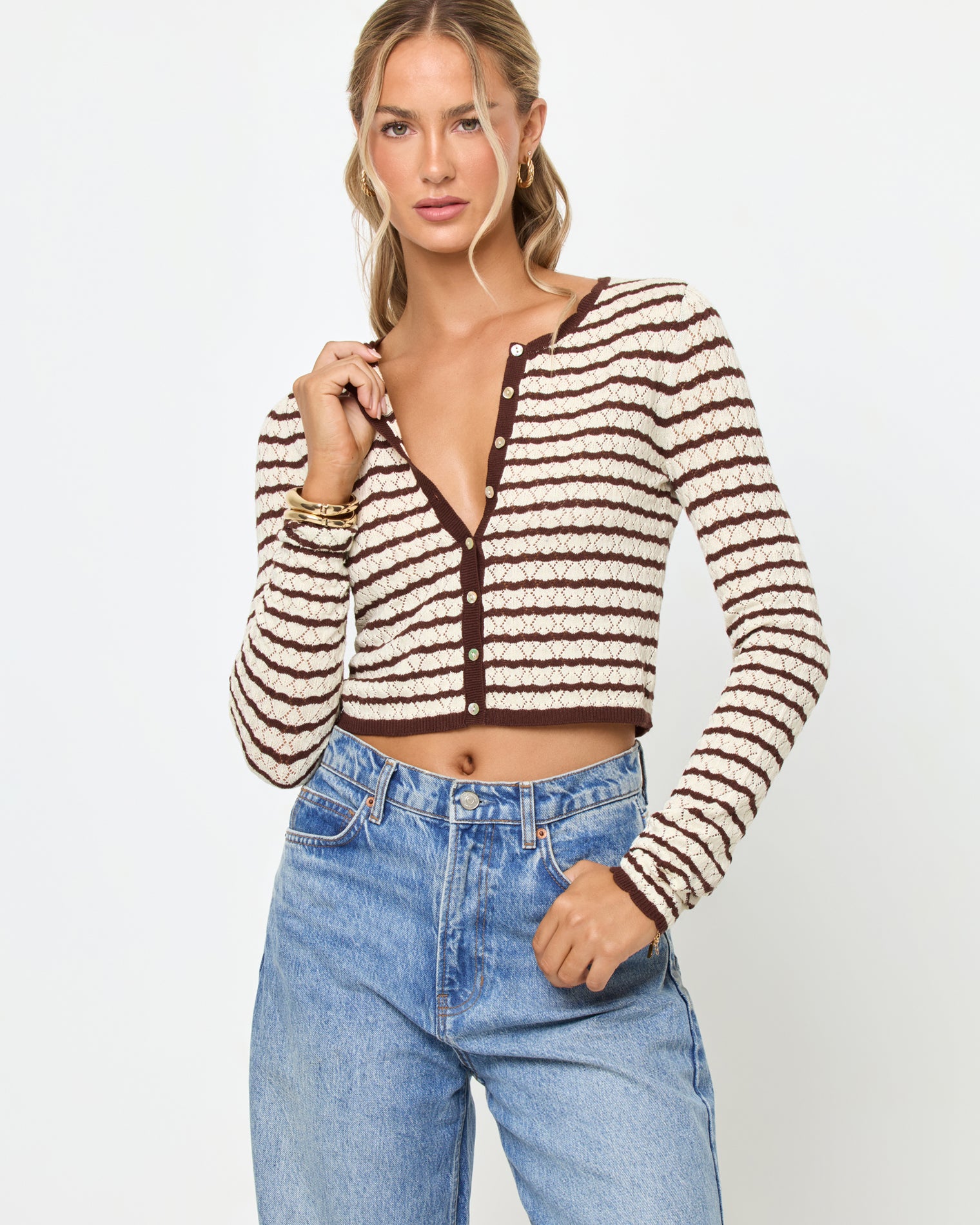 Rosalie Cardigan - Cafe-Cream Front view of a model wearing the LSPACE Rosalie Cardigan in cafe-cream, a cropped knit cardigan with cream and dark brown horizontal stripes, a V-neckline, and gold button closure, styled with light wash jeans.