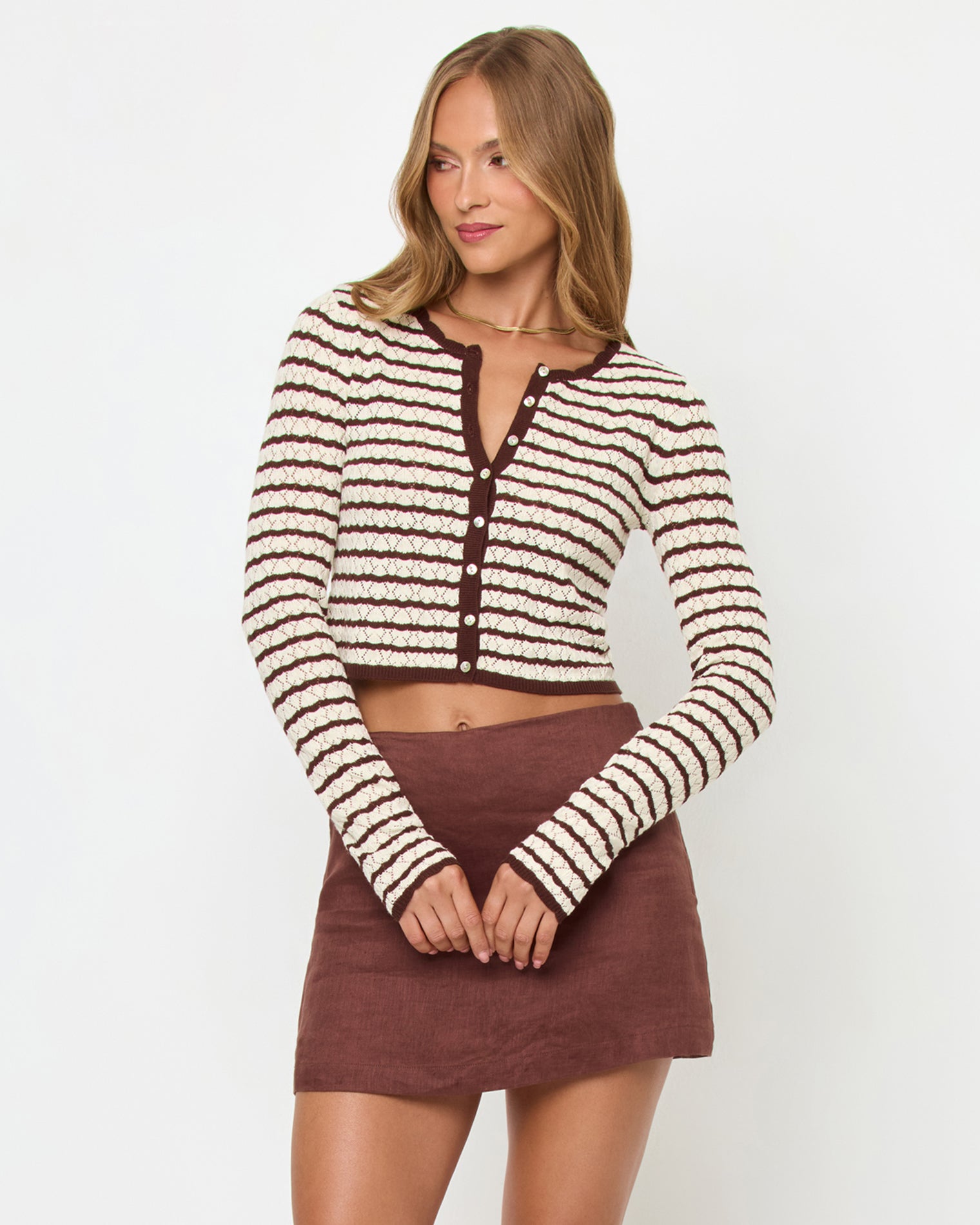 Rosalie Cardigan - Cafe-Cream Front view of a model wearing the LSPACE Rosalie Cardigan in cafe-cream, a cropped knit cardigan with cream and dark brown horizontal stripes, V-neckline, and gold button closure, styled with a brown mini skirt.