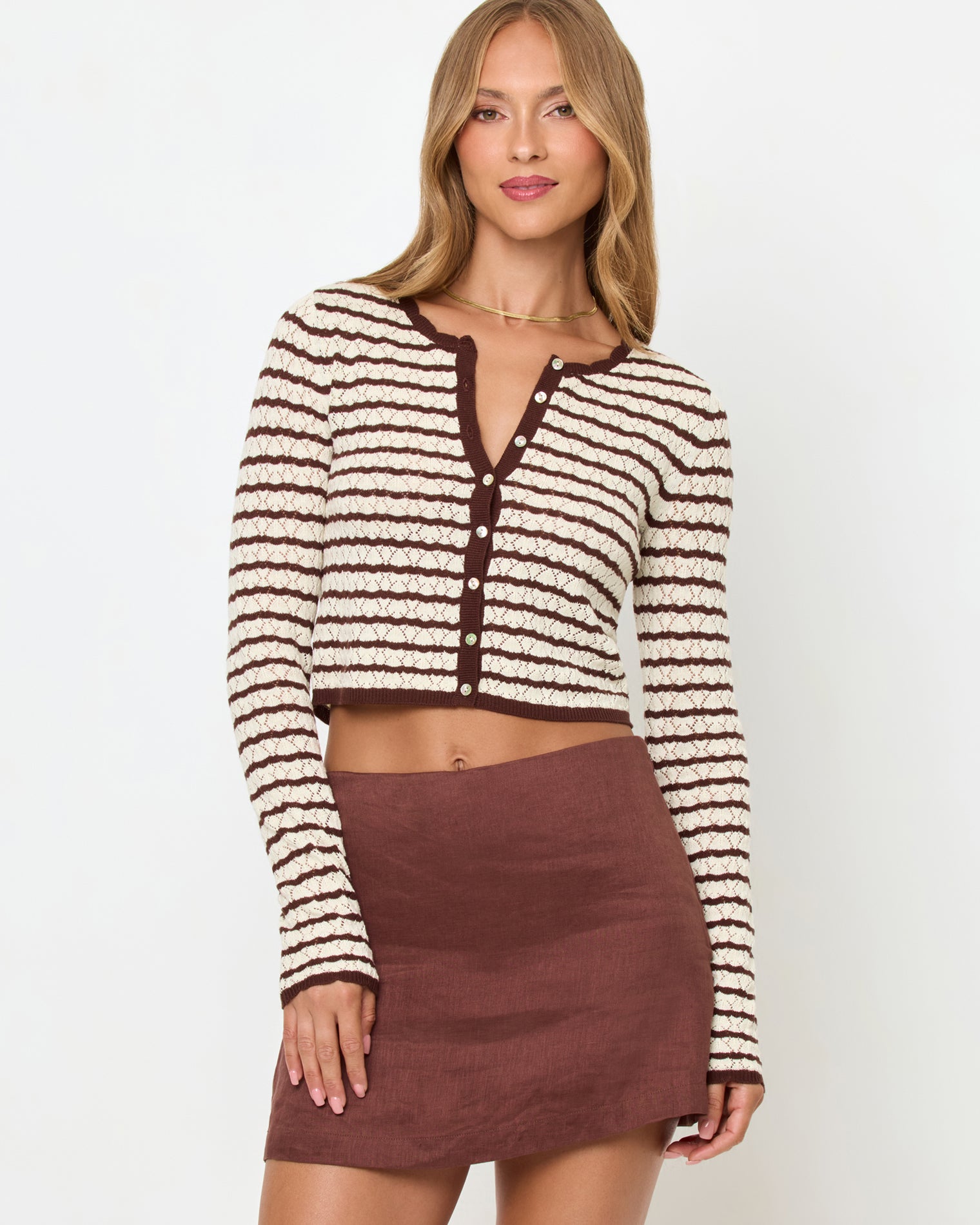 Rosalie Cardigan - Cafe-Cream Front view of a model wearing the LSPACE Rosalie Cardigan in cafe-cream, a cropped knit cardigan with cream and dark brown horizontal stripes, a V-neckline, and gold button closure, styled with a brown linen mini skirt.