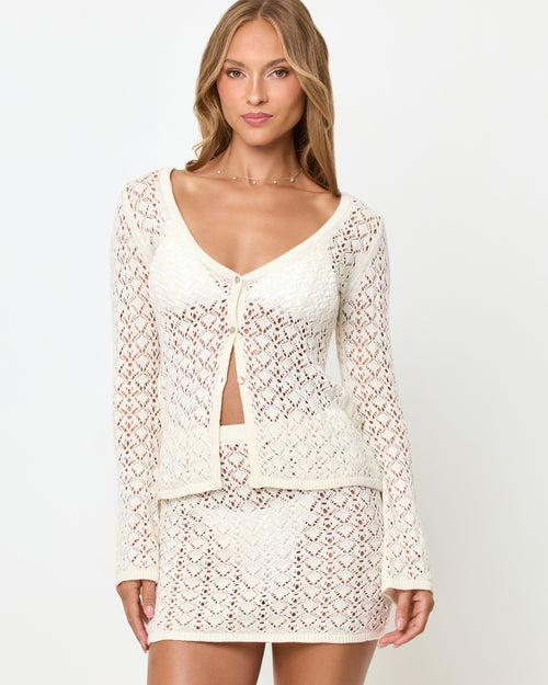 Three-quarter front view of a model wearing the LSPACE Nova Top in cream, a long-sleeve crochet top with a deep V-neckline and button tie-front closure, styled with the matching LSPACE Nova Skirt.