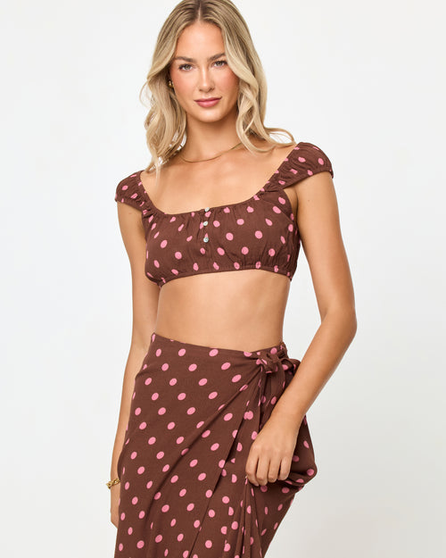 Front view of a model wearing the LSPACE Ever Top in alotta polka dot, a chocolate brown and pink polka dot crop top with cap sleeves and a square neckline, styled with the matching wrap skirt.
