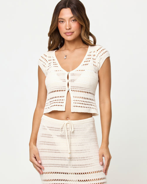 Front view of model wearing the LSPACE Tula Top in cream, a semi-sheer crochet cropped top with cap sleeves and button front, styled with matching cream crochet maxi skirt.