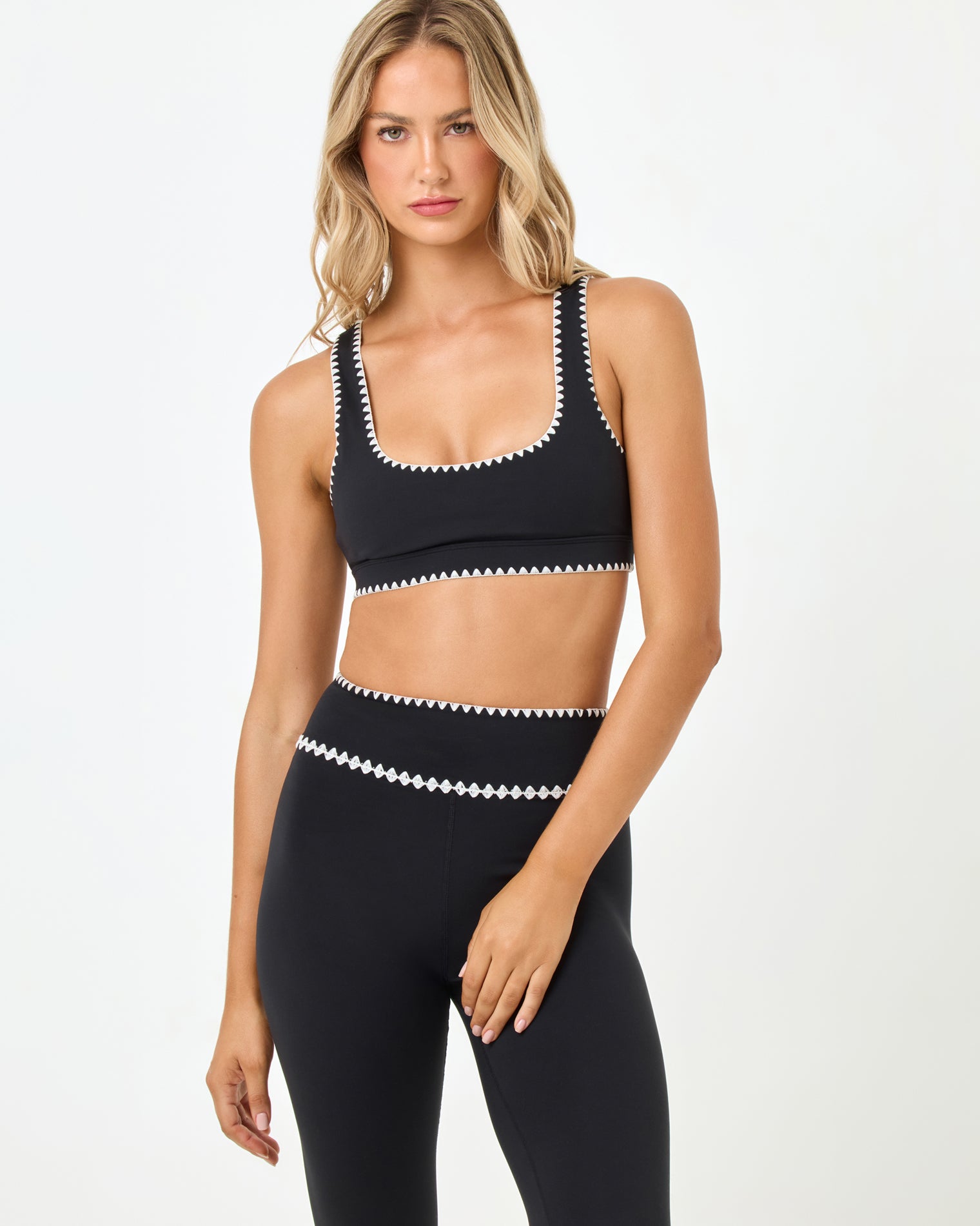 Heartbreaker Bra - Black Front studio three-quarter view of the Heartbreaker Bra in black, a black scoop-neck sports bra with white zig-zag diamond trim at the neckline, straps, and underbust band, styled with matching black high-waist leggings with white zig-zag trim at the waist.