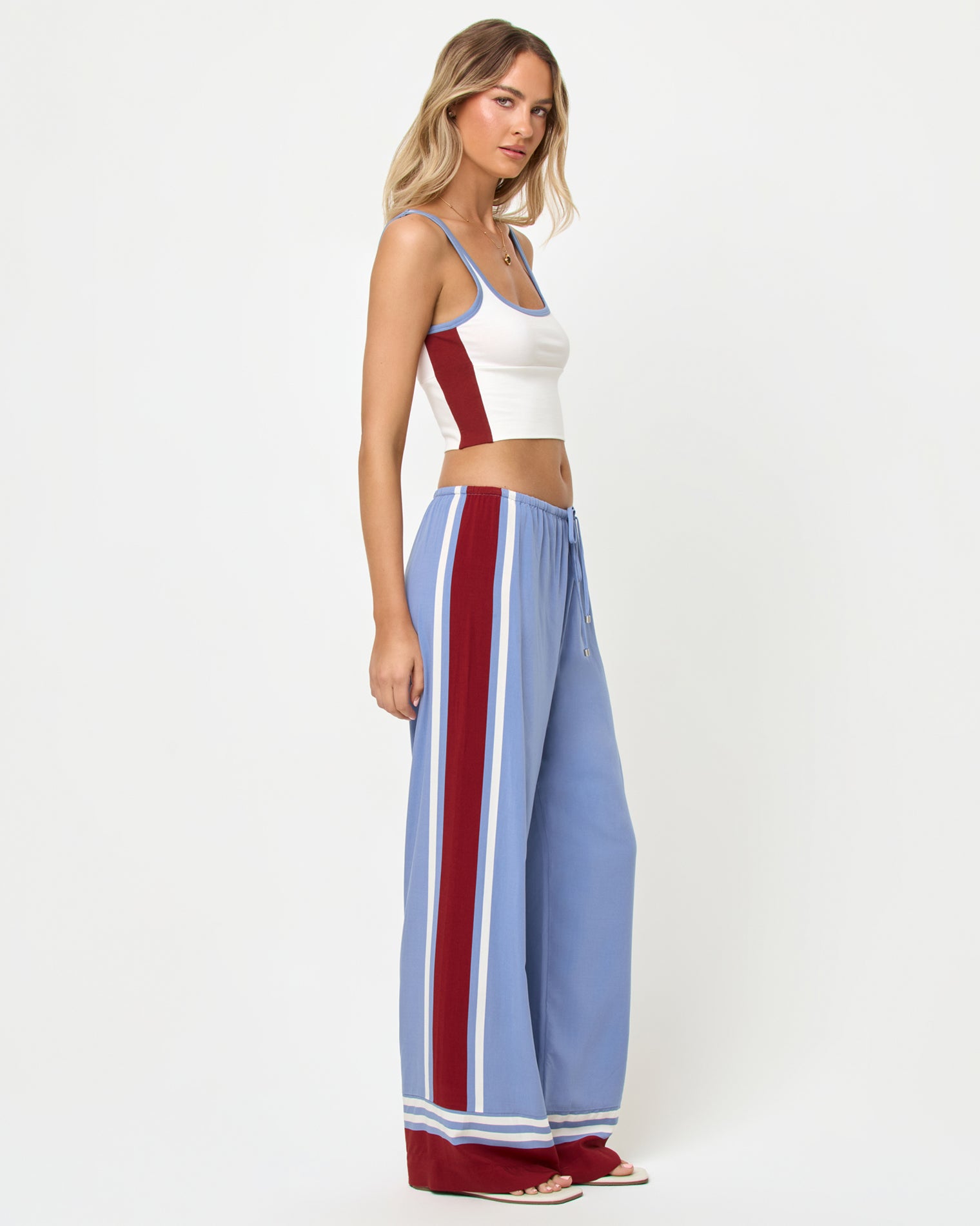 Unwind Pant - Unwind Stripe Three-quarter front side profile studio view of the Reset Tank and Unwind Pant in Cream-Chambray-Fig, a cream ribbed scoop-neck crop cami with chambray blue and burgundy trim and color-block sides paired with chambray blue wide-leg stripe pants with a wide burgundy vertical side stripe and cream piping, styled with nude heeled sandals.