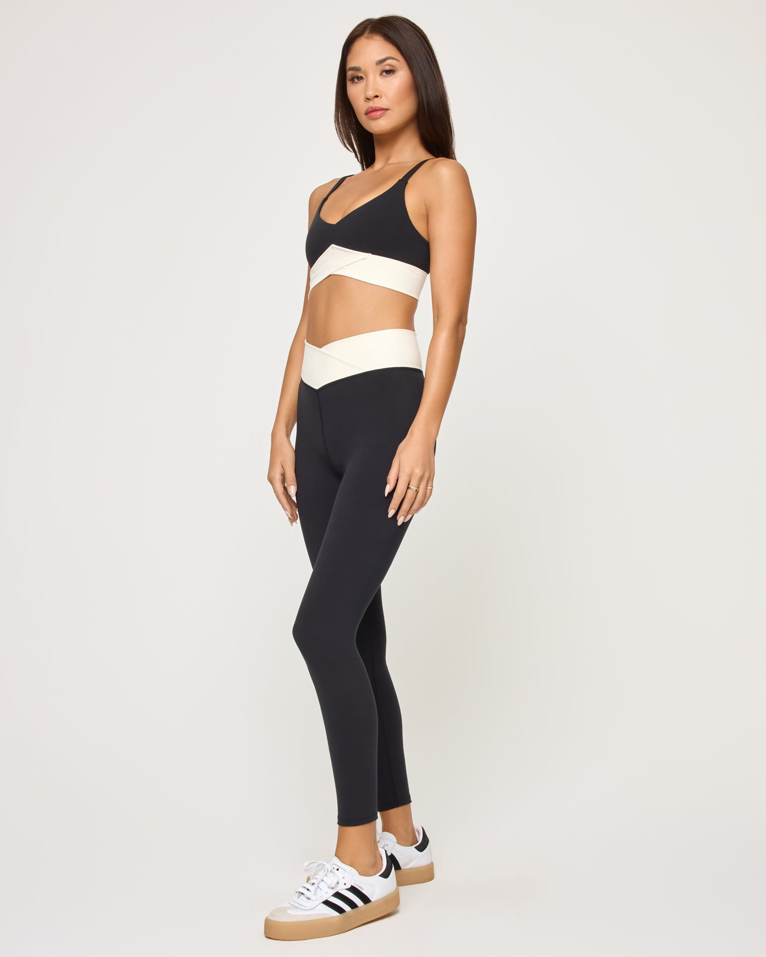 Unmatched Legging - Black-Cream Side view of the LSPACE Unmatched Legging in black-cream on a model against a white background, paired with the matching black crossover sports bra with cream band detail, and black and white sneakers.