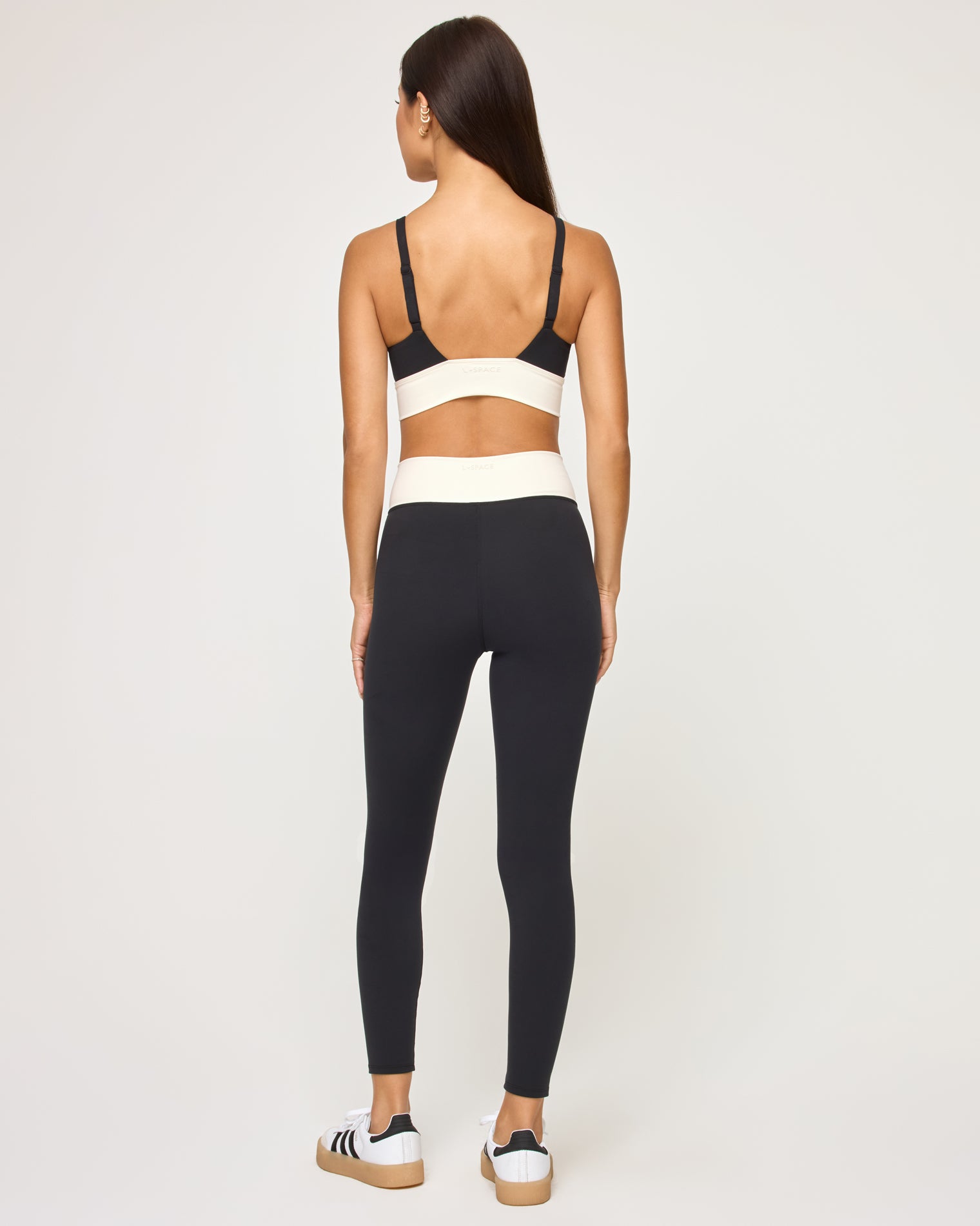 Unmatched Legging - Black-Cream Back view of the LSPACE Unmatched Legging in black-cream on a model against a white background, showing the cream crossover waistband detail and the open back of the matching black sports bra, with black and white sneakers.