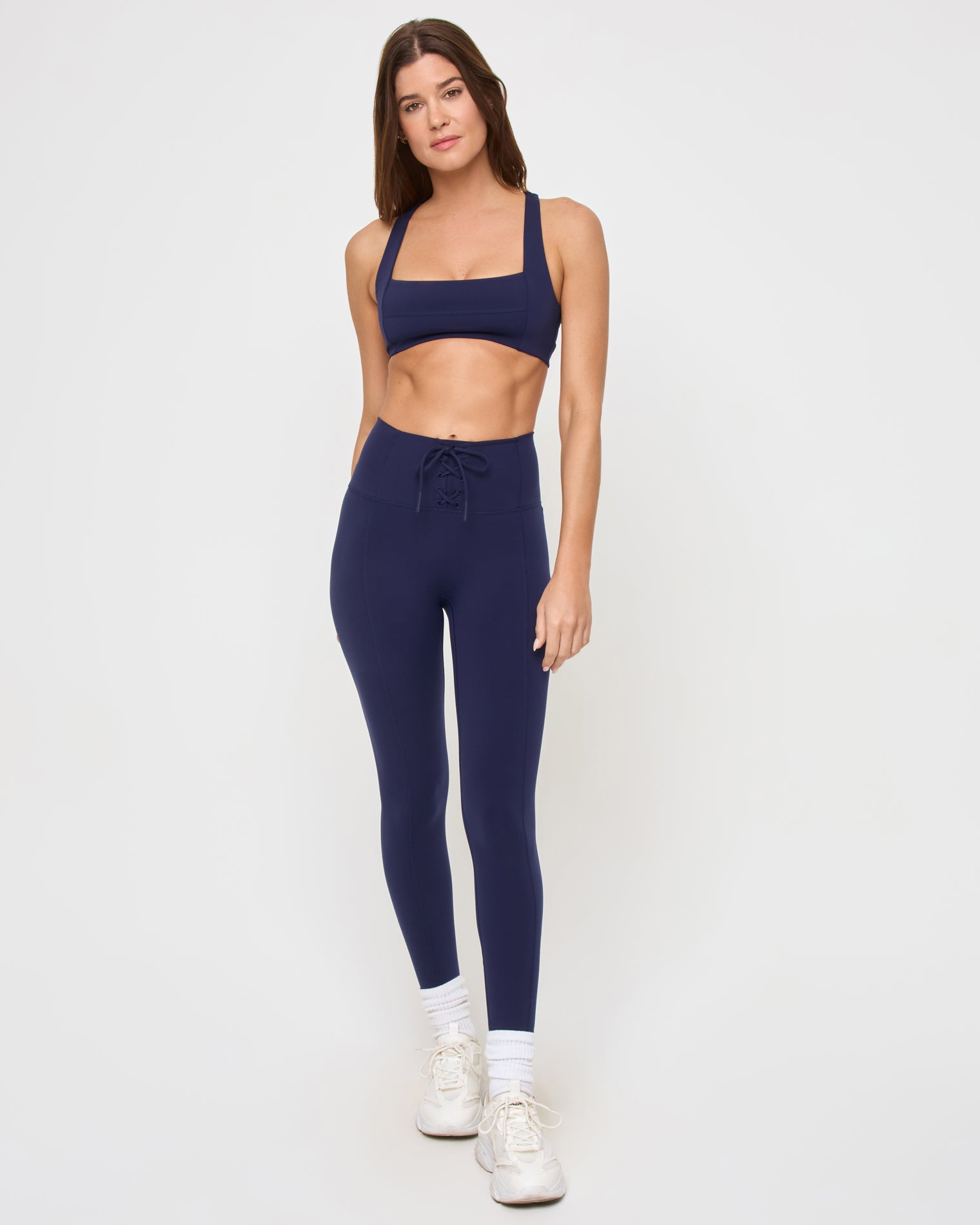 Uppercut Bra - Midnight Blue Front view of the LSPACE Uppercut Bra in midnight blue on a model against a white background, showing the full-length outfit with the midnight navy square-neck sports bra and matching high-rise full-length leggings or pants with lace-up waistband, white sneakers and socks.