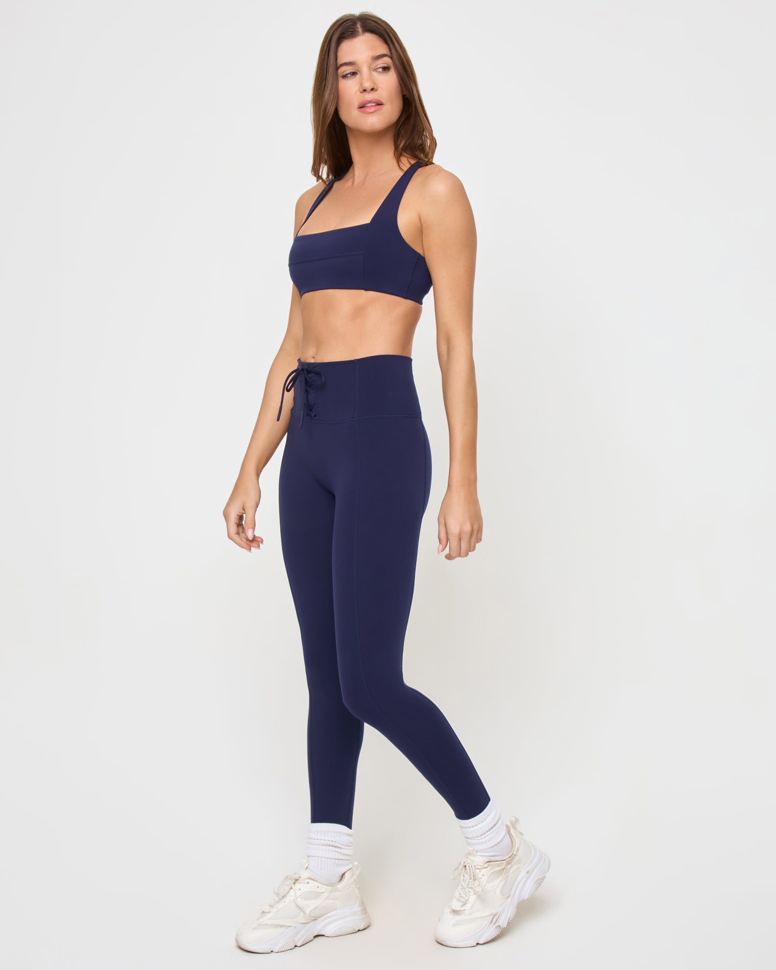Uppercut Bra - Midnight Blue Side view of the LSPACE Uppercut Bra in midnight blue on a model against a white background, showing the square-neck sports bra profile with the lace-up side waistband detail of the matching midnight blue active pants, white sneakers and socks.
