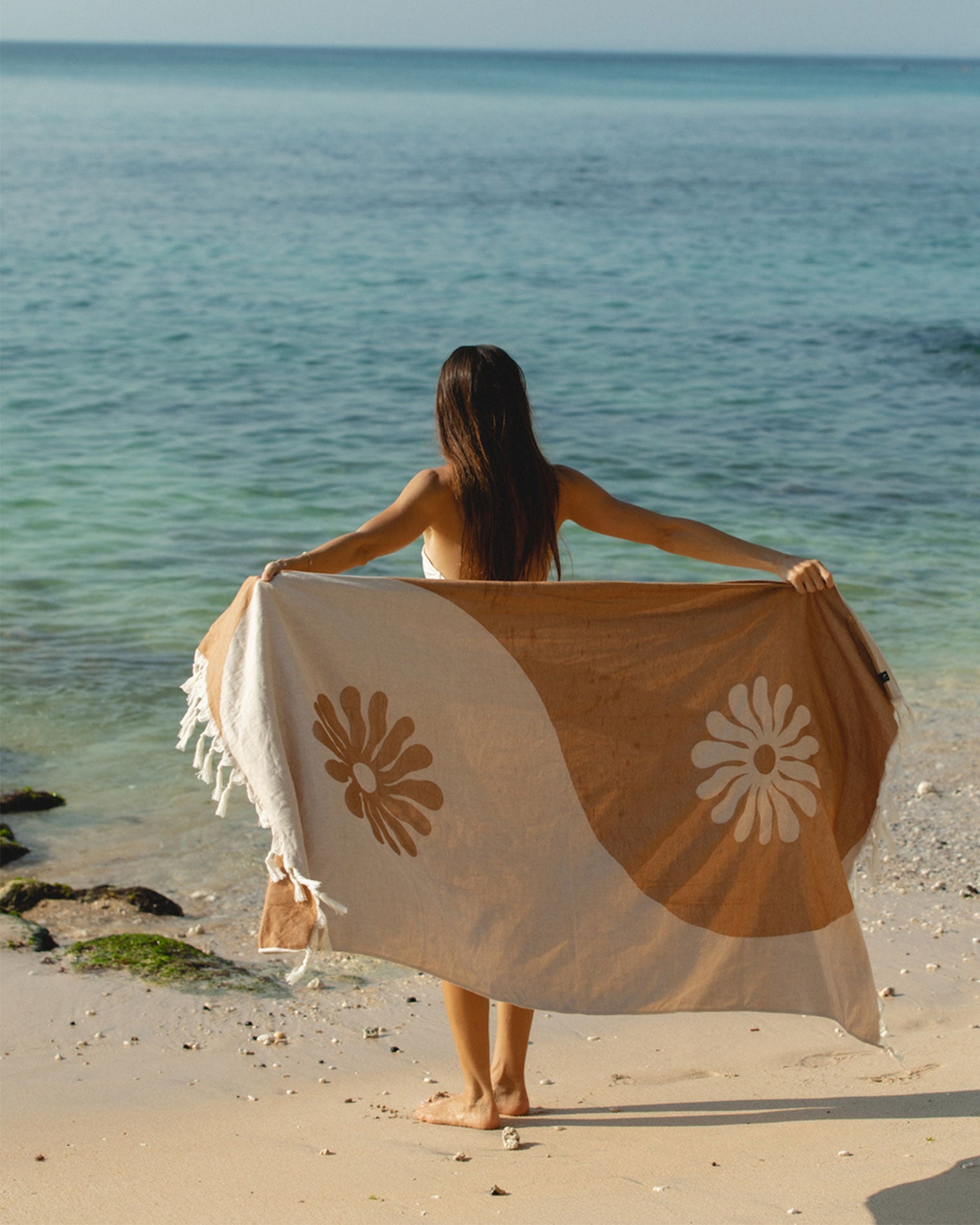 Slowtide Botanic Balance Turkish Towel - Brush Model on the beach holding the Slowtide Botanic Balance Turkish Towel in brush open behind her back, facing away toward the ocean, displaying the tan and cream botanical pattern.