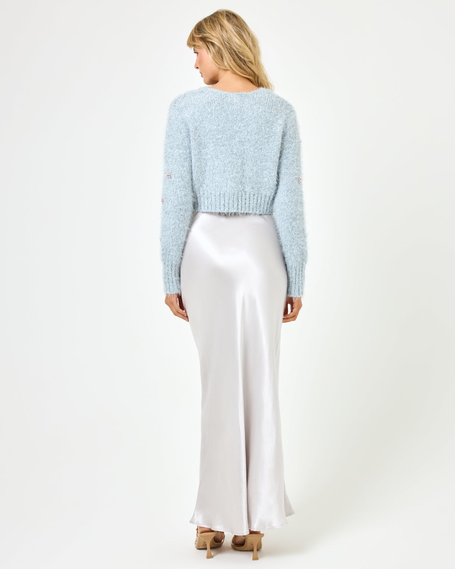 Crystal Cardigan - Dove Full body back view of a model wearing the LSPACE Crystal Cardigan in dove, paired with a silver satin maxi skirt, showing the back of the cropped fluffy knit cardigan with long sleeves and ribbed hem.