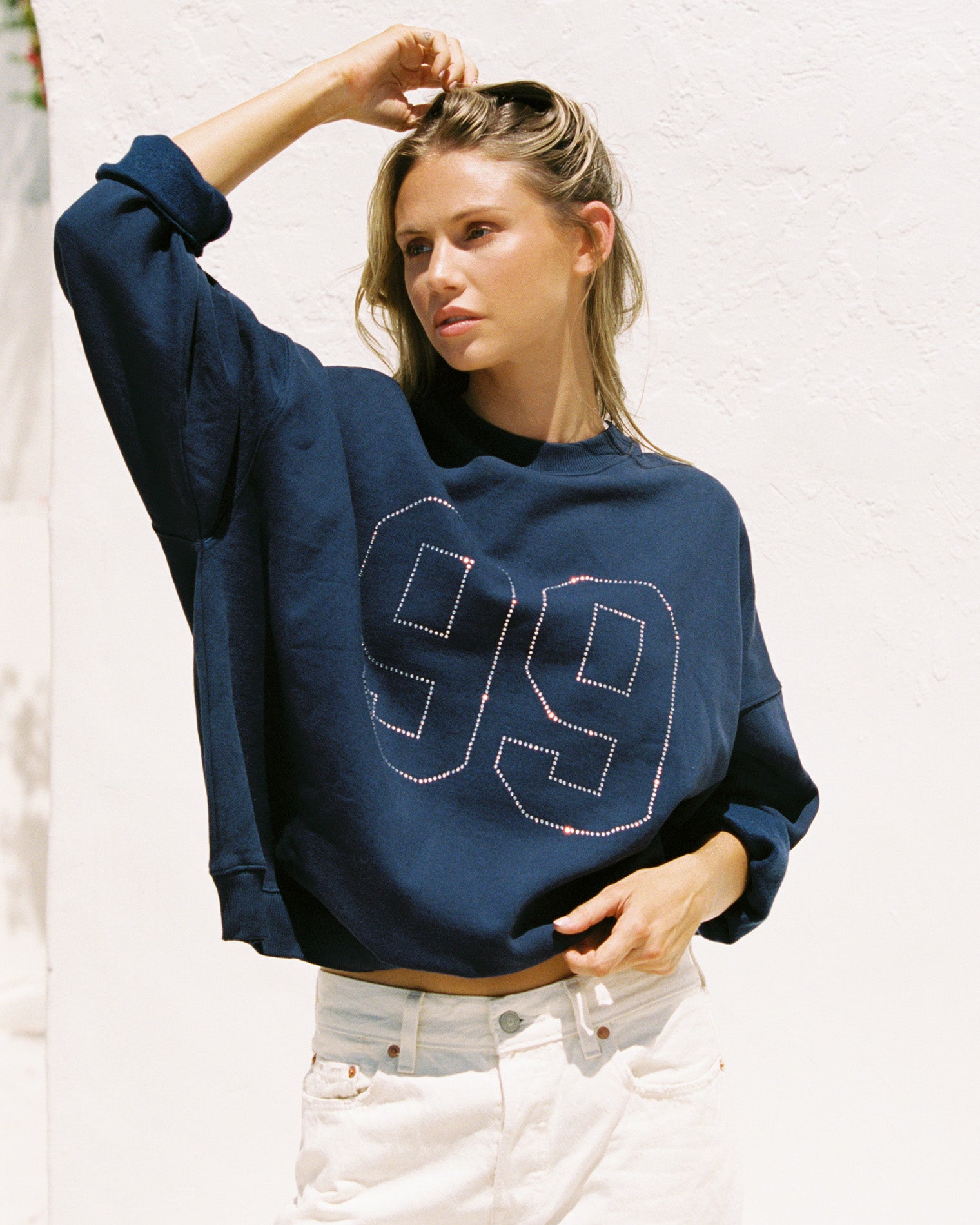 Legend Sweatshirt - Midnight Blue Lifestyle editorial shot of a model wearing the LSPACE Legend Sweatshirt in midnight blue, an oversized cropped sweatshirt with a rhinestone number graphic, paired with white denim jeans against an outdoor white wall.