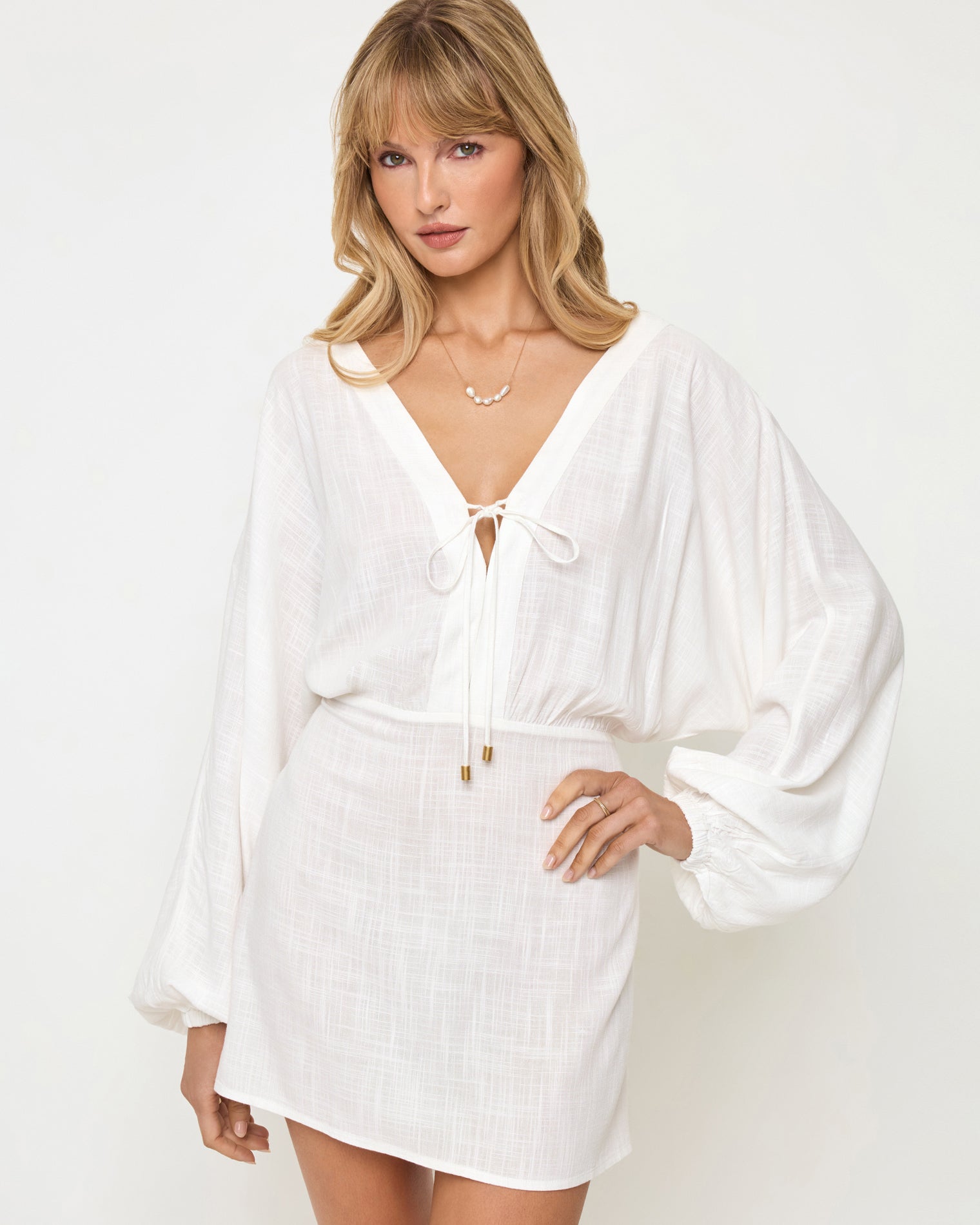 Deia Mini Dress - Cream Close-up of a model wearing the LSPACE Deia Mini Dress in cream, showing the deep V-neckline with tie-front closure and gold hardware, wide balloon sleeves, and elasticized waistband in cream textured linen fabric.
