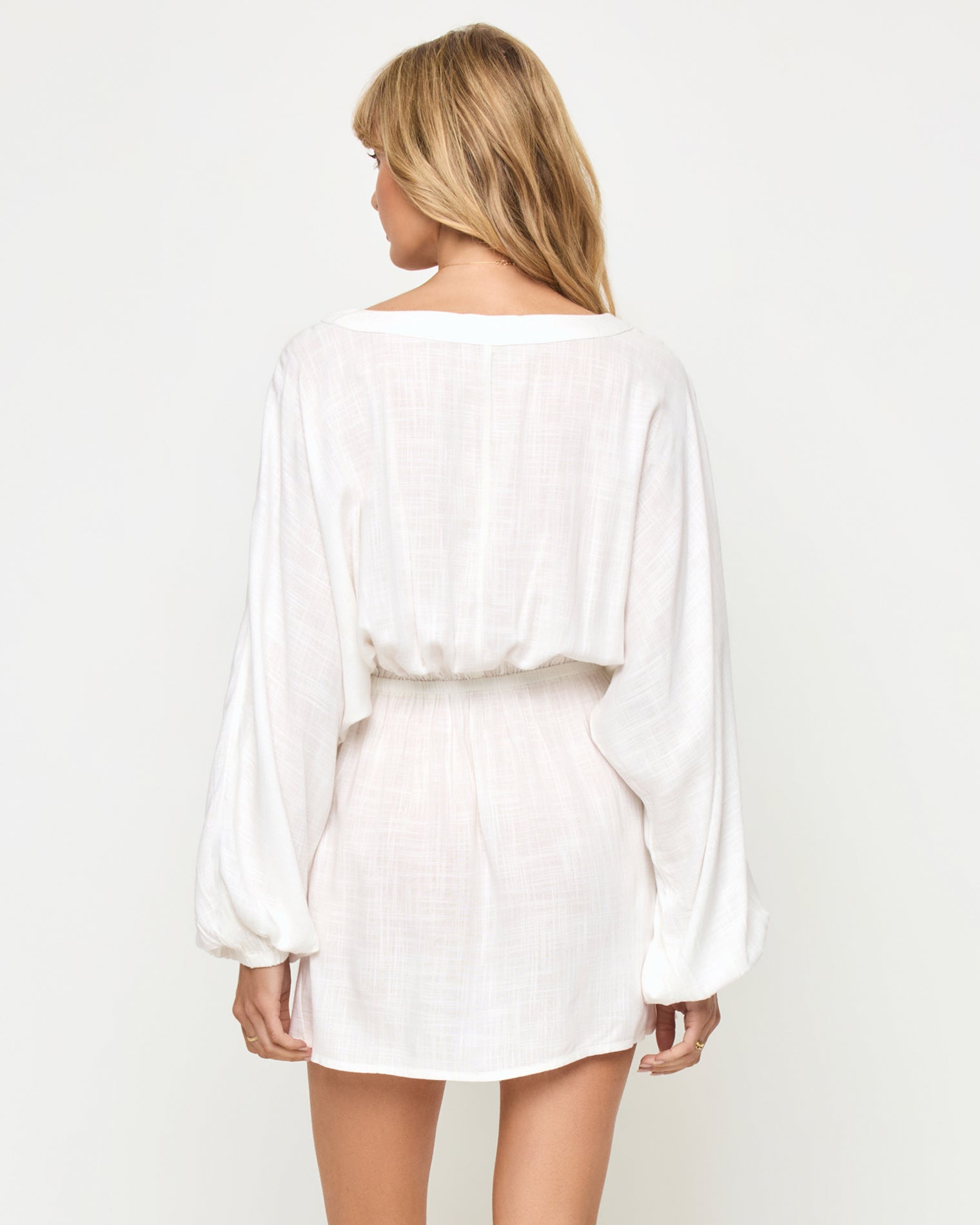 Deia Mini Dress - Cream Back view of a model wearing the LSPACE Deia Mini Dress in cream, showing the open V-back, wide balloon sleeves, elastic waistband, and mini length in cream linen fabric.