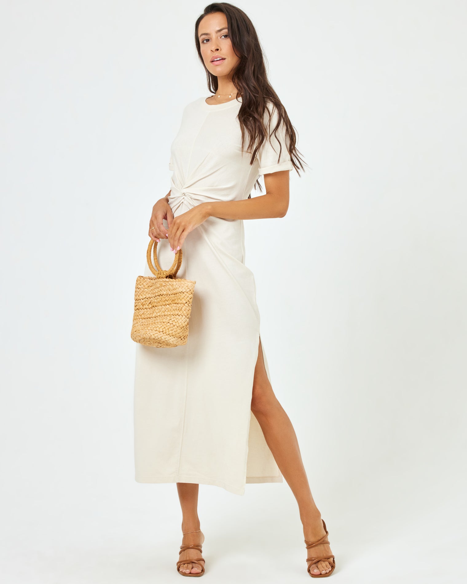 Drew Dress - Cream Angled front view of a model wearing the LSPACE Drew Dress in cream, a short-sleeve jersey midi dress with a twisted knot at the waist, styled with a woven tote bag.