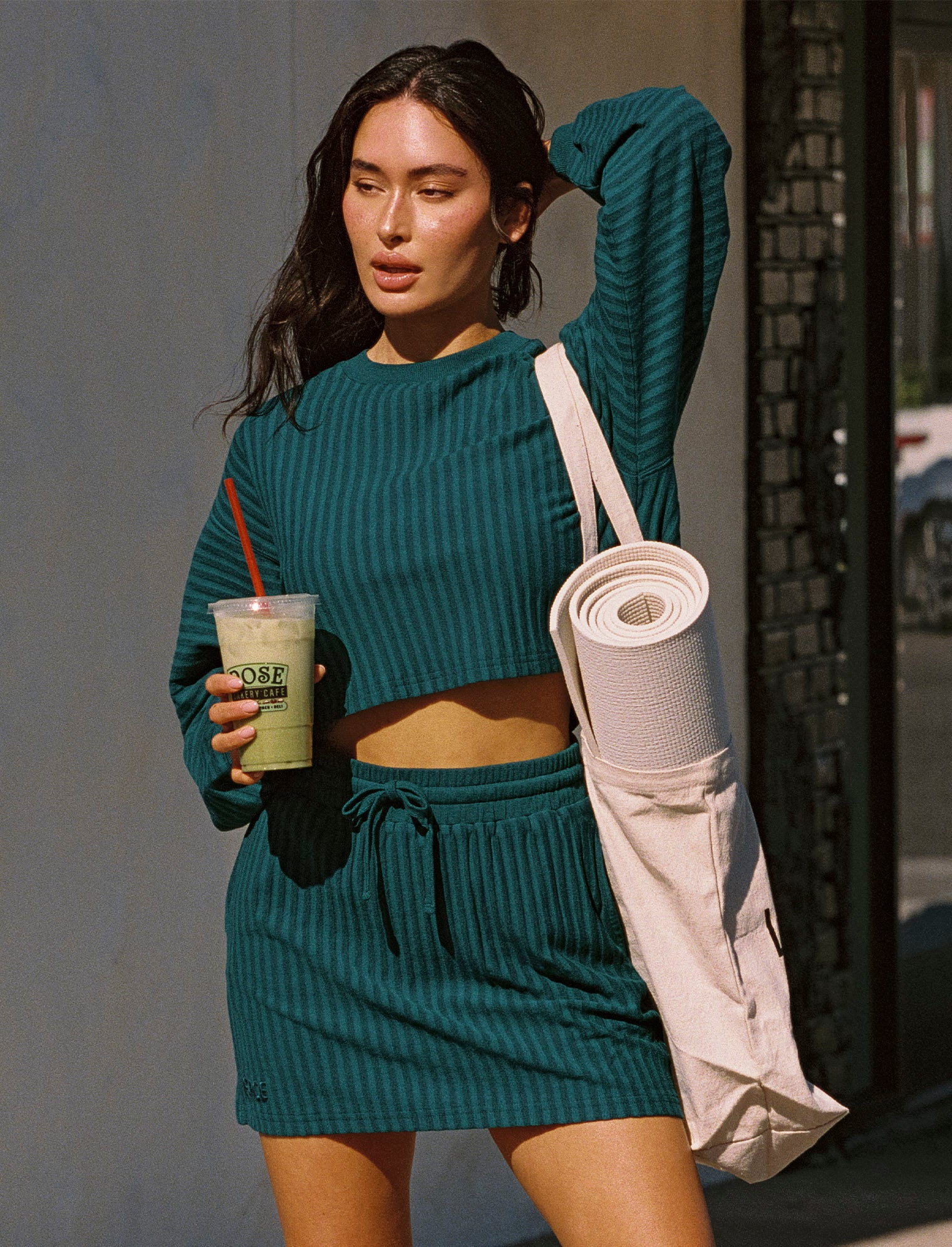 Half Court Pullover - Deep Teal Model outdoors on a city street wearing the Half Court Pullover in deep teal, a ribbed long-sleeve cropped crewneck, with matching deep teal Half Court Skirt, holding a green drink and carrying a yoga mat bag, Adidas sneakers.