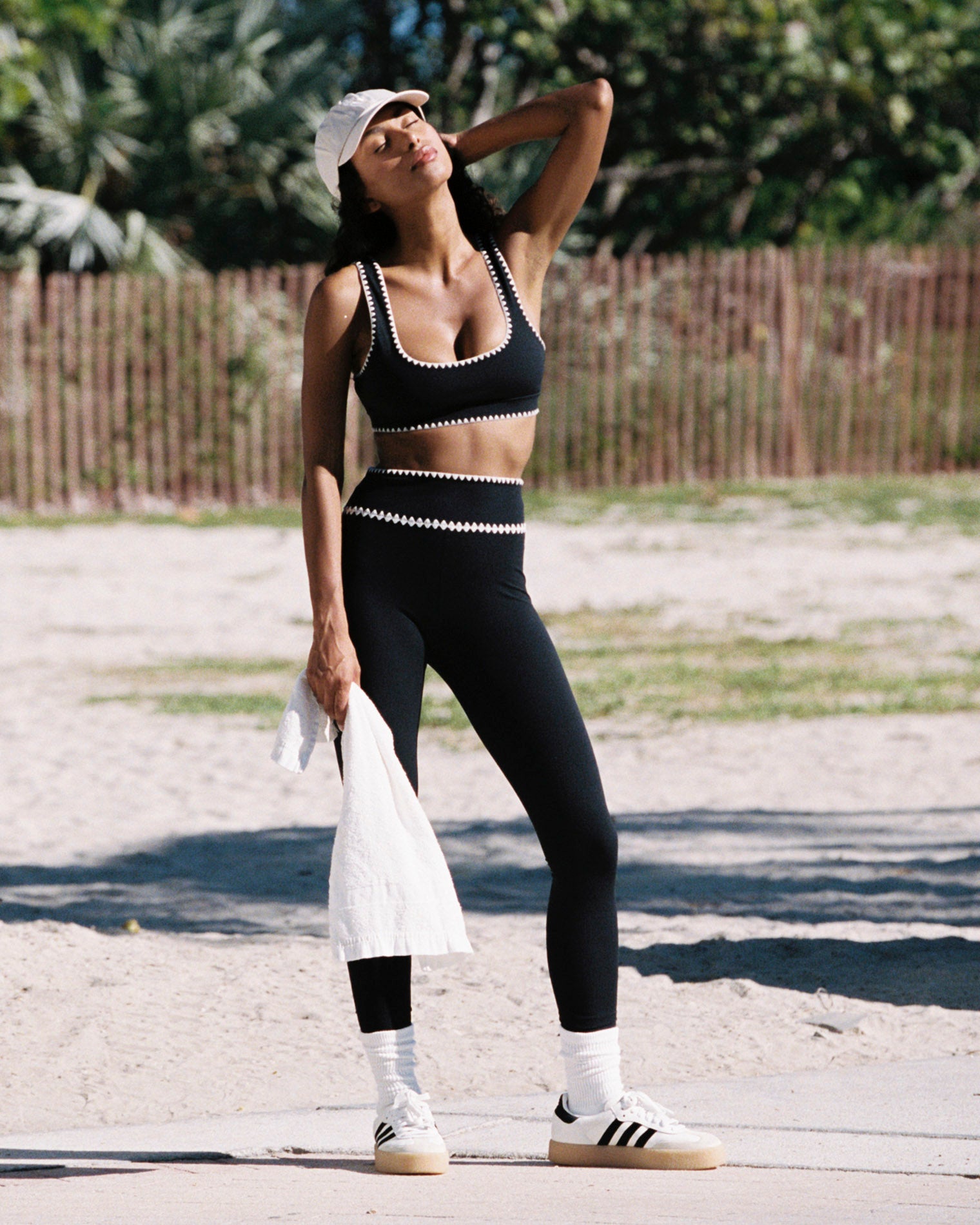 Heartbreaker Legging - Black Outdoor lifestyle front full-length view of the Heartbreaker Legging in black, black high-waist leggings with white zig-zag diamond trim at the waist, styled with a matching black sports bra, white baseball cap, white Adidas sneakers with white crew socks, holding a white jacket, against a beach and palm tree background.