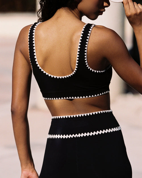 Outdoor lifestyle back and torso close-up of the Heartbreaker Bra in black, a black sports bra with white zig-zag diamond trim along the straps and underbust band, worn with matching black high-waist leggings with white zig-zag trim at the waist.