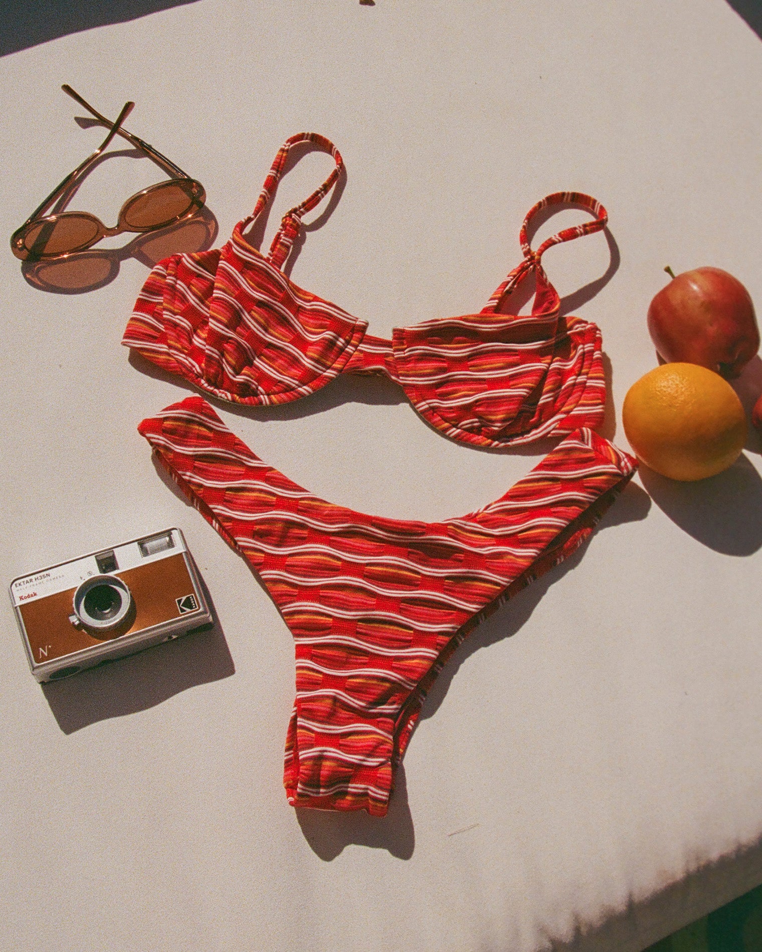x The Salty Blonde Rocky Bikini Bottom - Tutti Frutti Off-model flat lay of the LSPACE x The Salty Blonde Tutti Frutti Rocky Bikini Bottom styled with sunglasses, a camera, and fruit.