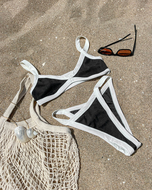 Off-model flat lay of the LSPACE black-cream Mackenzie Bikini Bottom and matching Taylor Bikini Top arranged on a sandy beach with sunglasses and a mesh beach bag nearby.