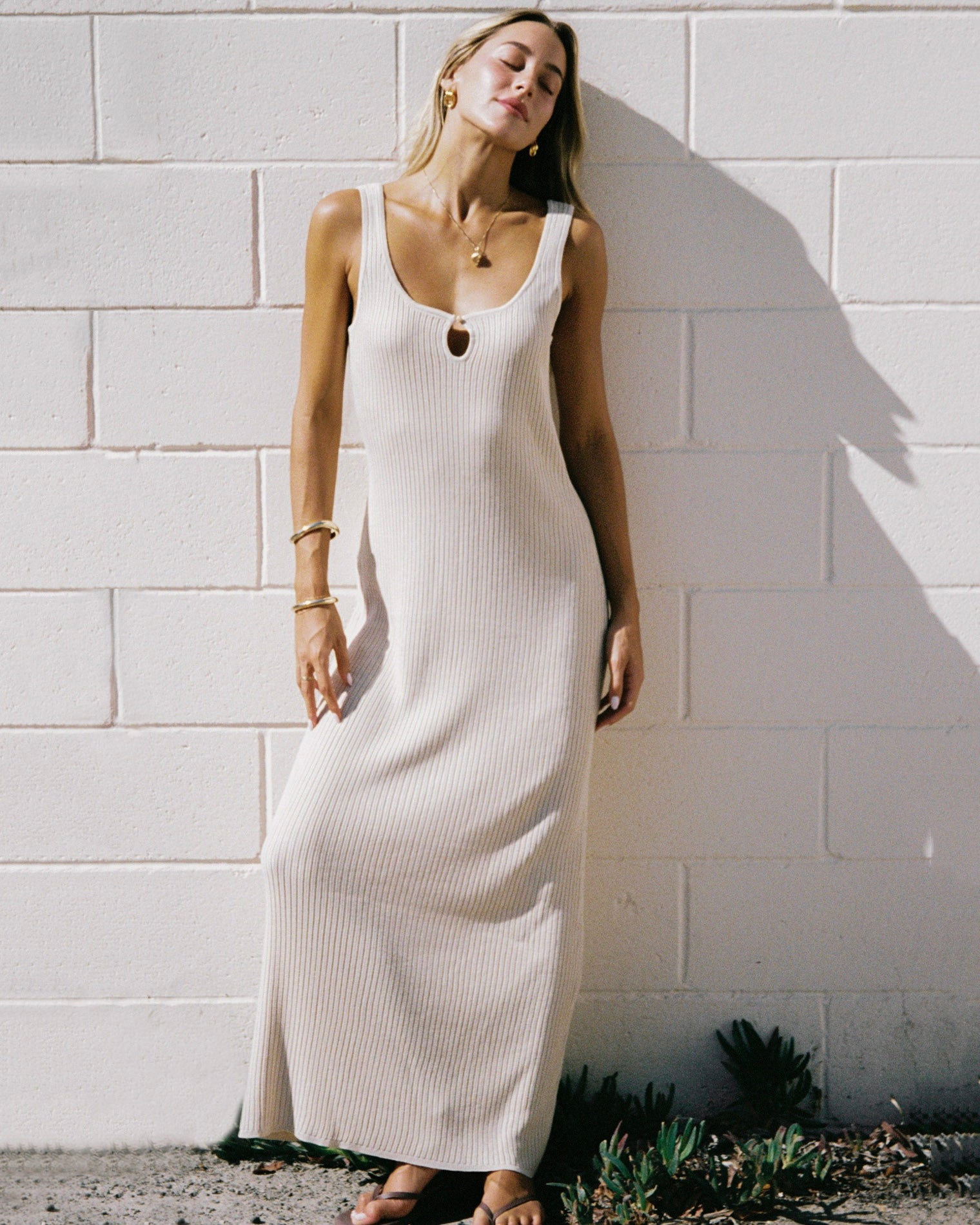 En Route Dress - Oat Editorial front view of a model leaning against a white brick wall, wearing the LSPACE En Route Dress - Oat in oat, a ribbed knit sleeveless maxi dress with a keyhole neckline.