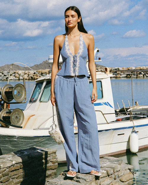 Front view on model of the LSPACE Sur Pant in Shell, wide-leg shell blue pants with a white crochet side trim, styled with a matching blue halter top and white sandals at a harbor.