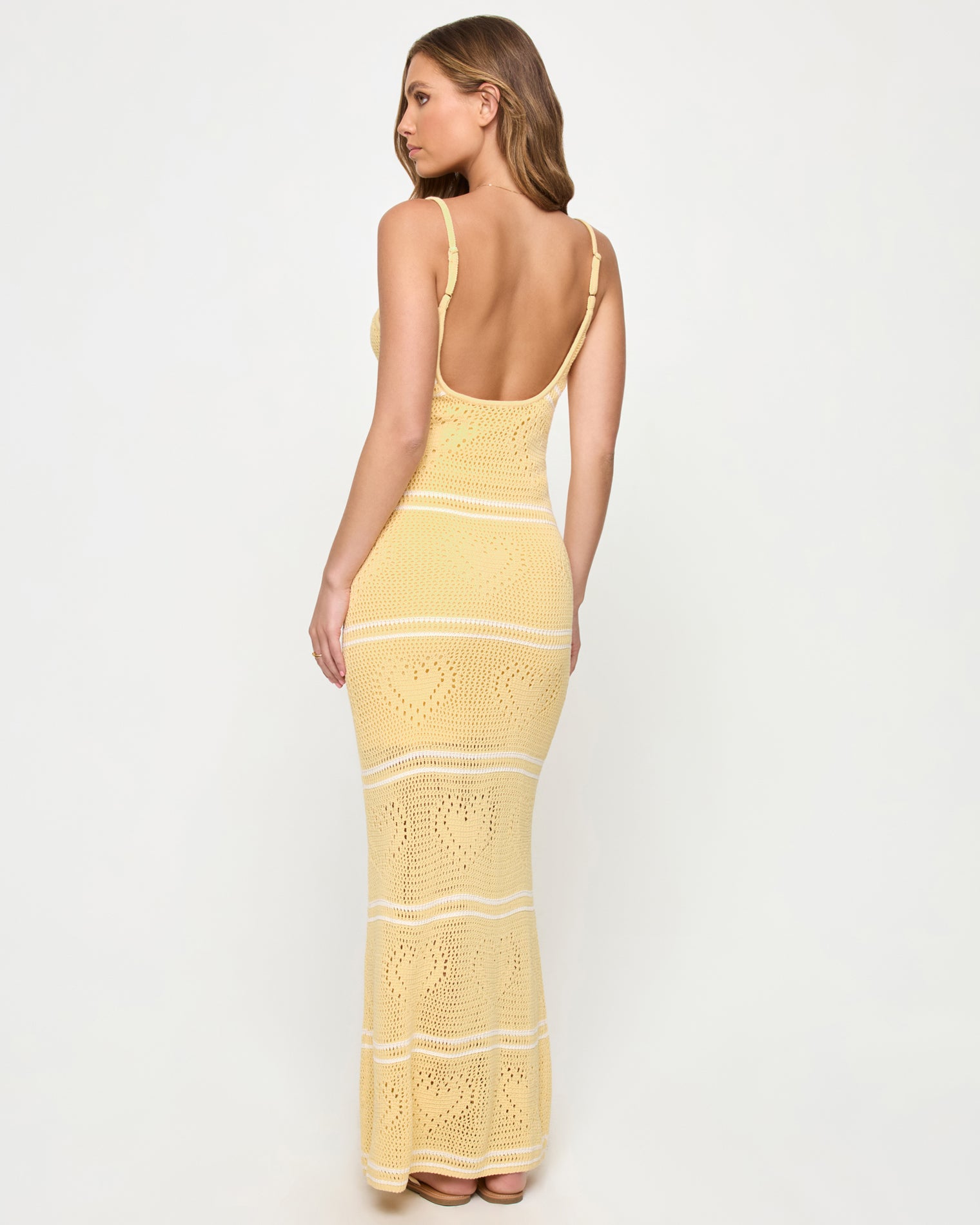Heart To Heart Dress - Daisy-Cream Back view of a model wearing the LSPACE Heart To Heart Dress in daisy-cream, showing the open low scoop back, adjustable spaghetti straps, and body-hugging crochet maxi silhouette in soft yellow with horizontal cream stripe details and heart motifs throughout.