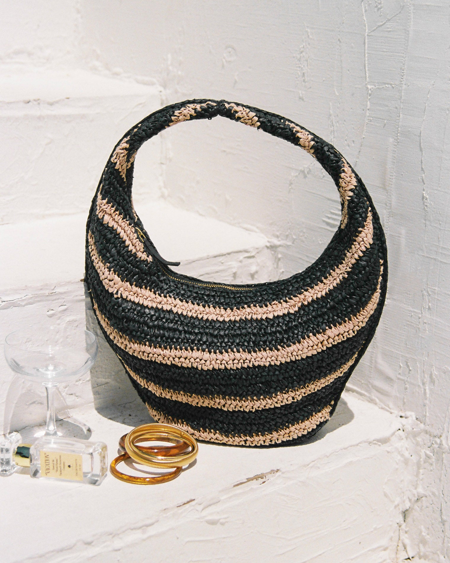 Aya Bag - Natural-Black Lifestyle image of the Aya Bag in natural-black, a crescent-shaped woven raffia hobo bag with black and natural horizontal stripes, styled on a white shelf with bangles and a perfume bottle.