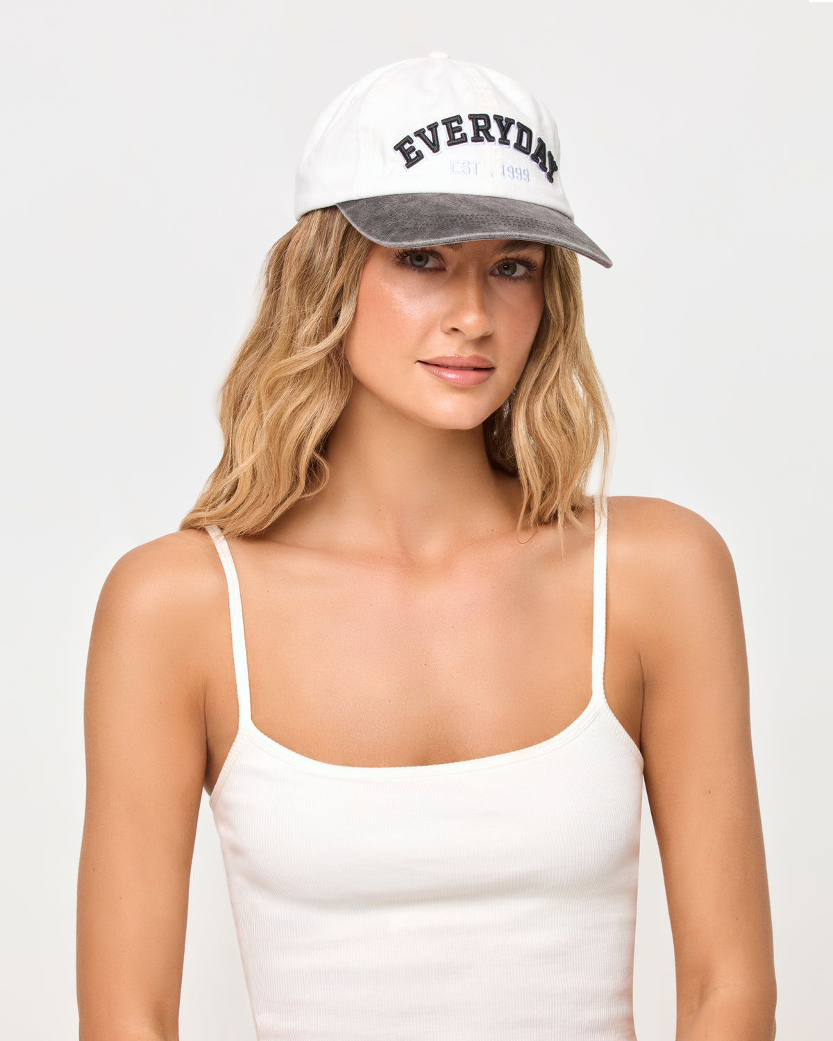 Buns Out Hat - Everyday A woman wearing a white baseball cap with a gray brim and 'EVERYDAY' printed in black on the front, paired with a white spaghetti-strap camisole top, against a light gray studio background.