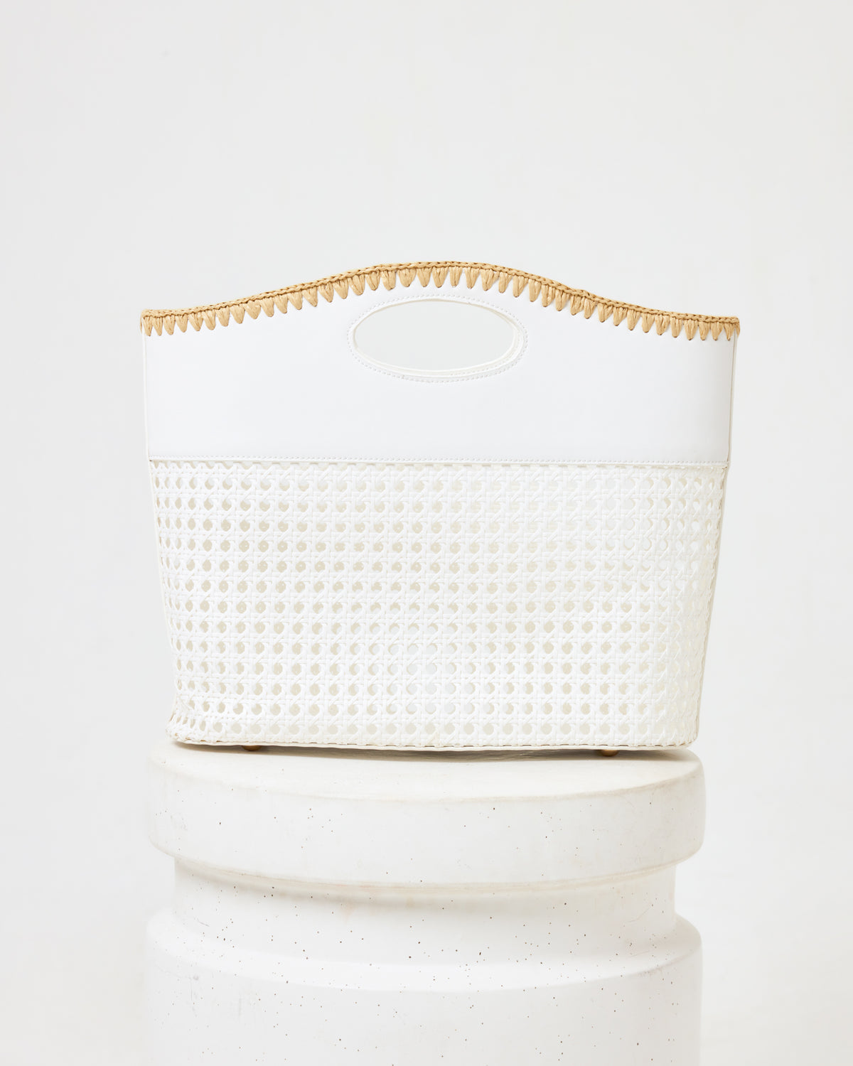 Off-model image of the Casablanca Bag in cream, a structured bag with a rattan weave bottom, cream fabric top, oval cutout handle, and natural trim, on a white pedestal.