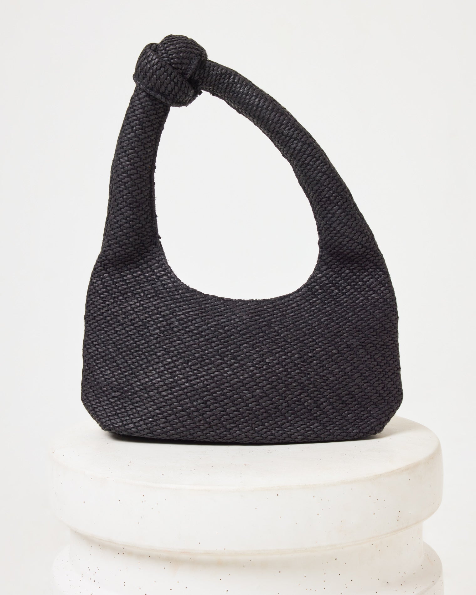 Ryder Bag - Black Off-model image of the Ryder Bag in black, a woven crescent hobo bag with a knotted handle, on a white pedestal.