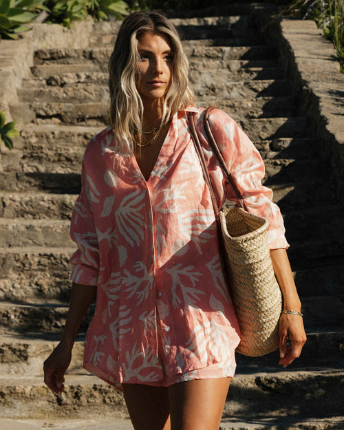 Front view of a model wearing the LSPACE Rio Tunic in shell or high water, a coral linen button-up shirt with a white tropical leaf print, styled with the matching Rio Short while walking down stone steps.