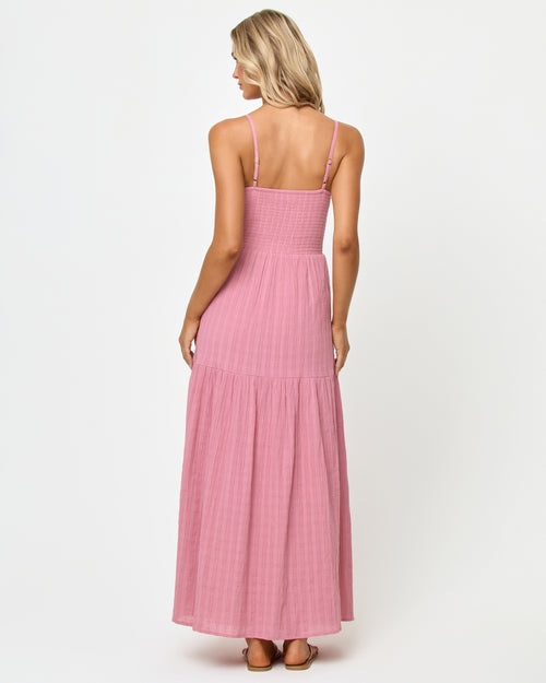Back view of a model wearing the LSPACE Mallorca Dress in cherry blossom, showing the smocked back bodice and tiered maxi skirt in a textured mauve-pink fabric.