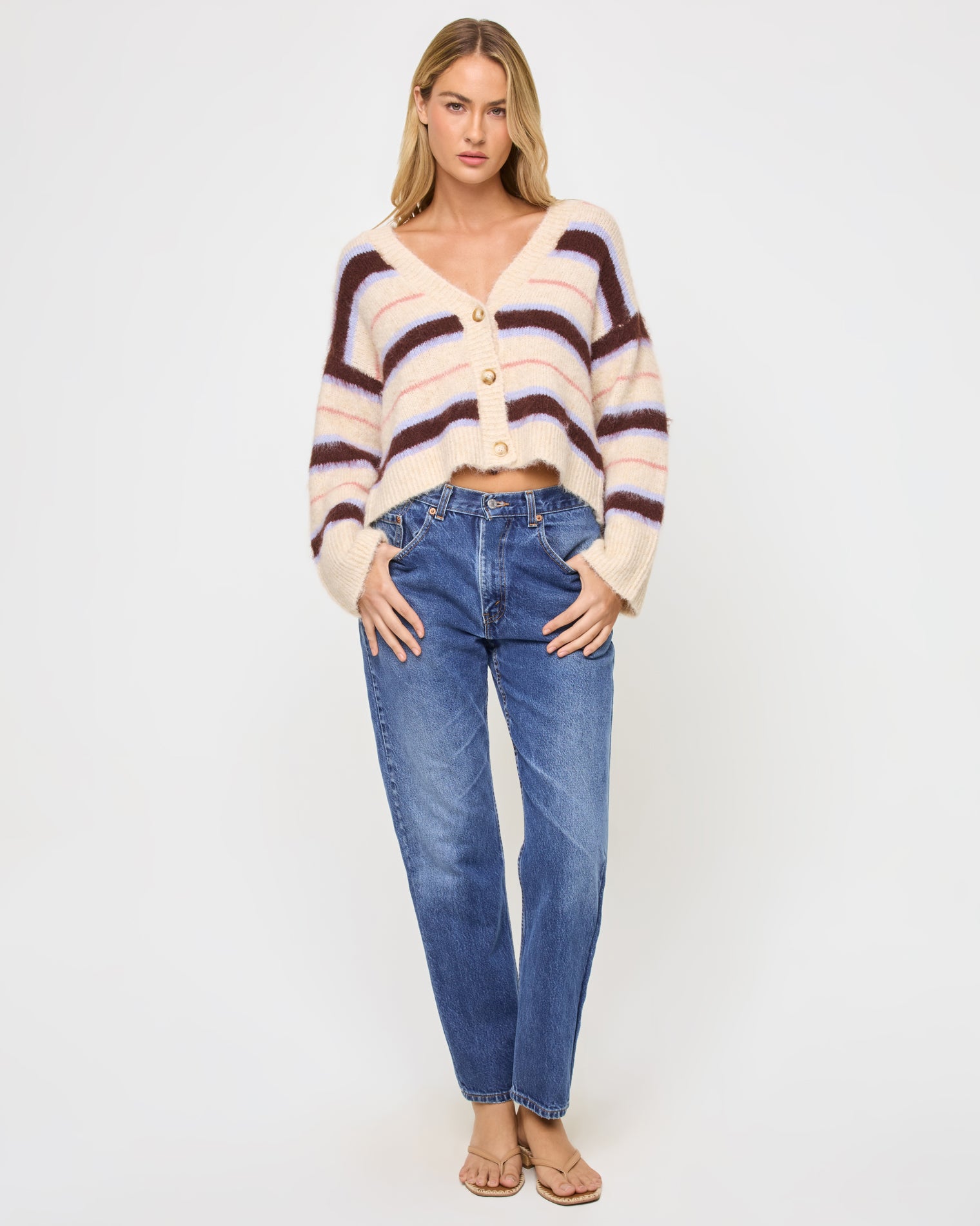 Montauk Cardigan - Prep School Stripe Full-length front view of a model wearing the LSPACE Montauk Cardigan in prep school stripe, a cropped oversized V-neck button-front cardigan in a soft eyelash knit with horizontal stripes in cream, dark brown, lavender, and pink, styled with jeans and sandals.