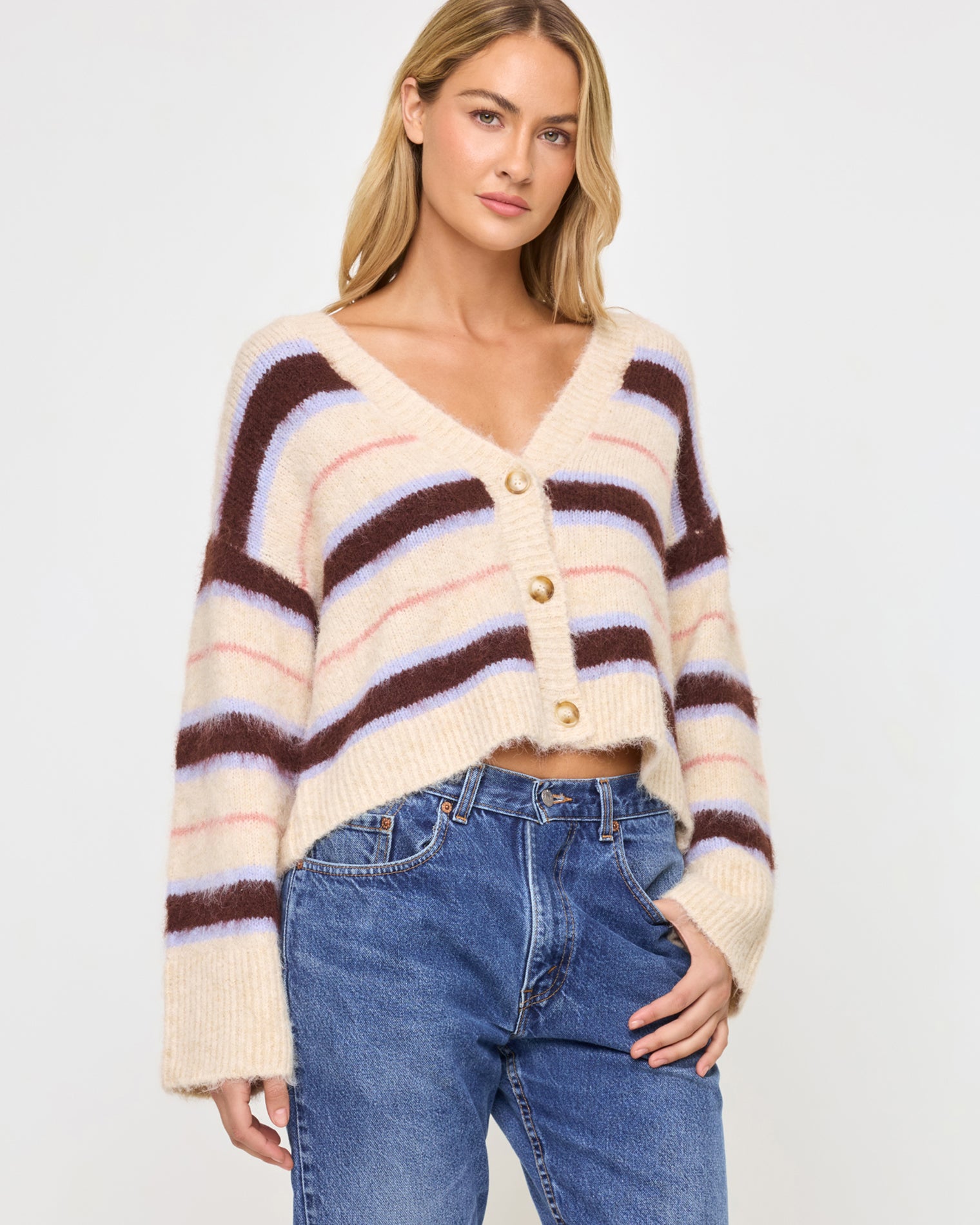 Montauk Cardigan - Prep School Stripe Front view of a model wearing the LSPACE Montauk Cardigan in prep school stripe, a cropped oversized V-neck button-front cardigan in a soft eyelash knit with horizontal stripes in cream, dark brown, lavender, and pink, styled with jeans.
