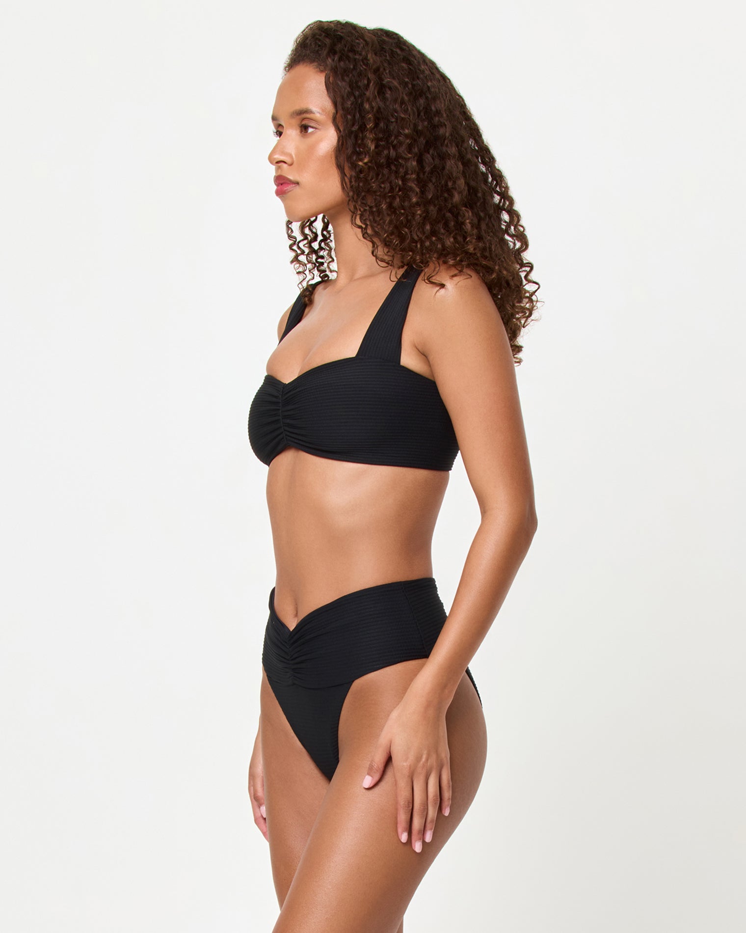 Marlee Bikini Top - Black Side view of a model wearing the LSPACE black Bardot Bikini Bottom, a ribbed high-waist style with gathered waistband detail, paired with the black Marlee Bikini Top.