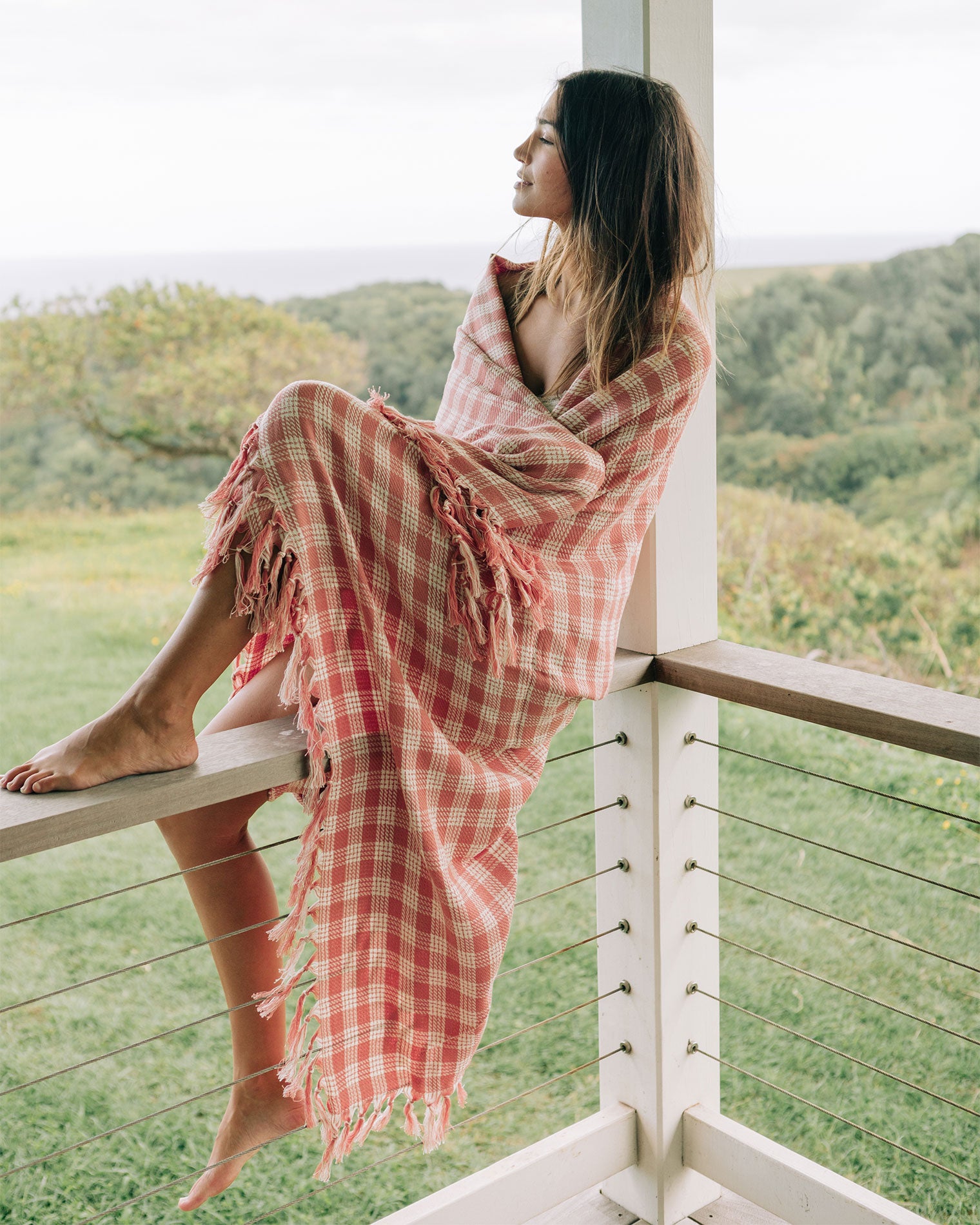 Slowtide Palaka Lightweight Beach Blanket - Guava Model wrapped in the Slowtide Palaka Lightweight Beach Blanket in guava while seated on a balcony railing overlooking greenery.