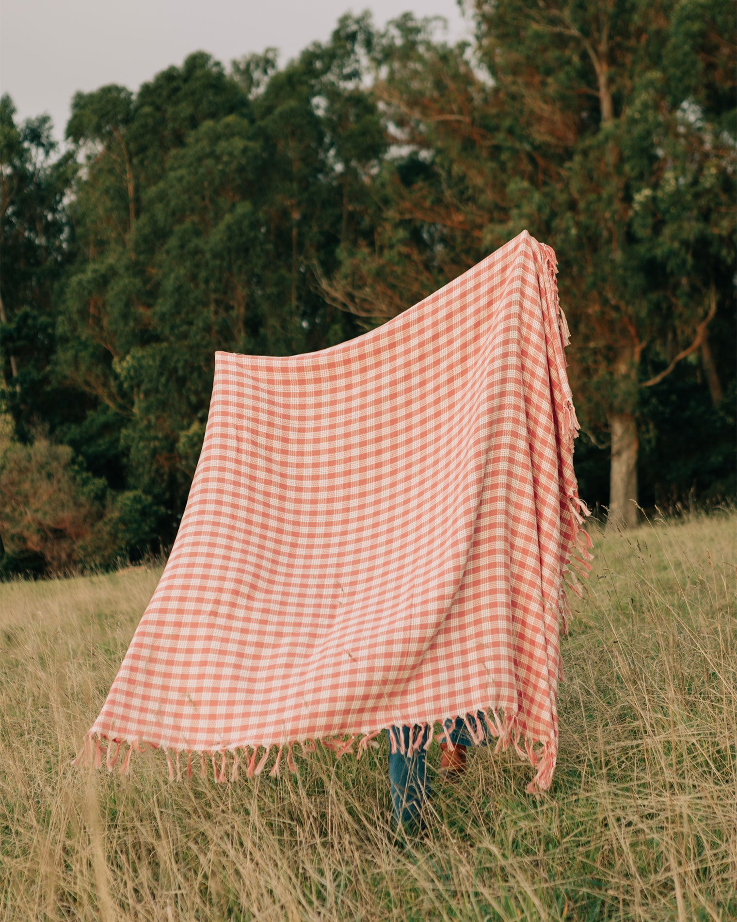 Slowtide Palaka Lightweight Beach Blanket - Guava The Slowtide Palaka Lightweight Beach Blanket in guava held up outdoors in a field with trees in the background.
