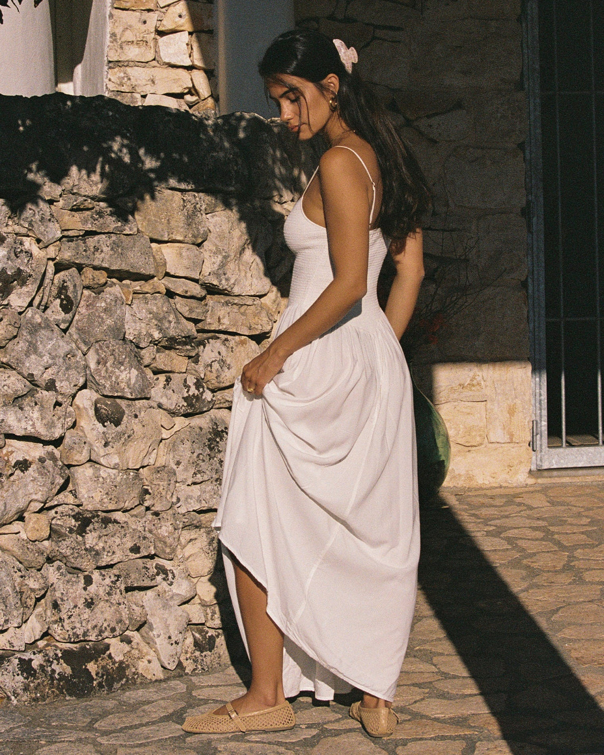 Misha Midi - Cream Lifestyle view of a model wearing the LSPACE Misha Midi in cream against a stone wall outdoors, featuring thin spaghetti straps, a smocked bodice, and a full flowing cream skirt.