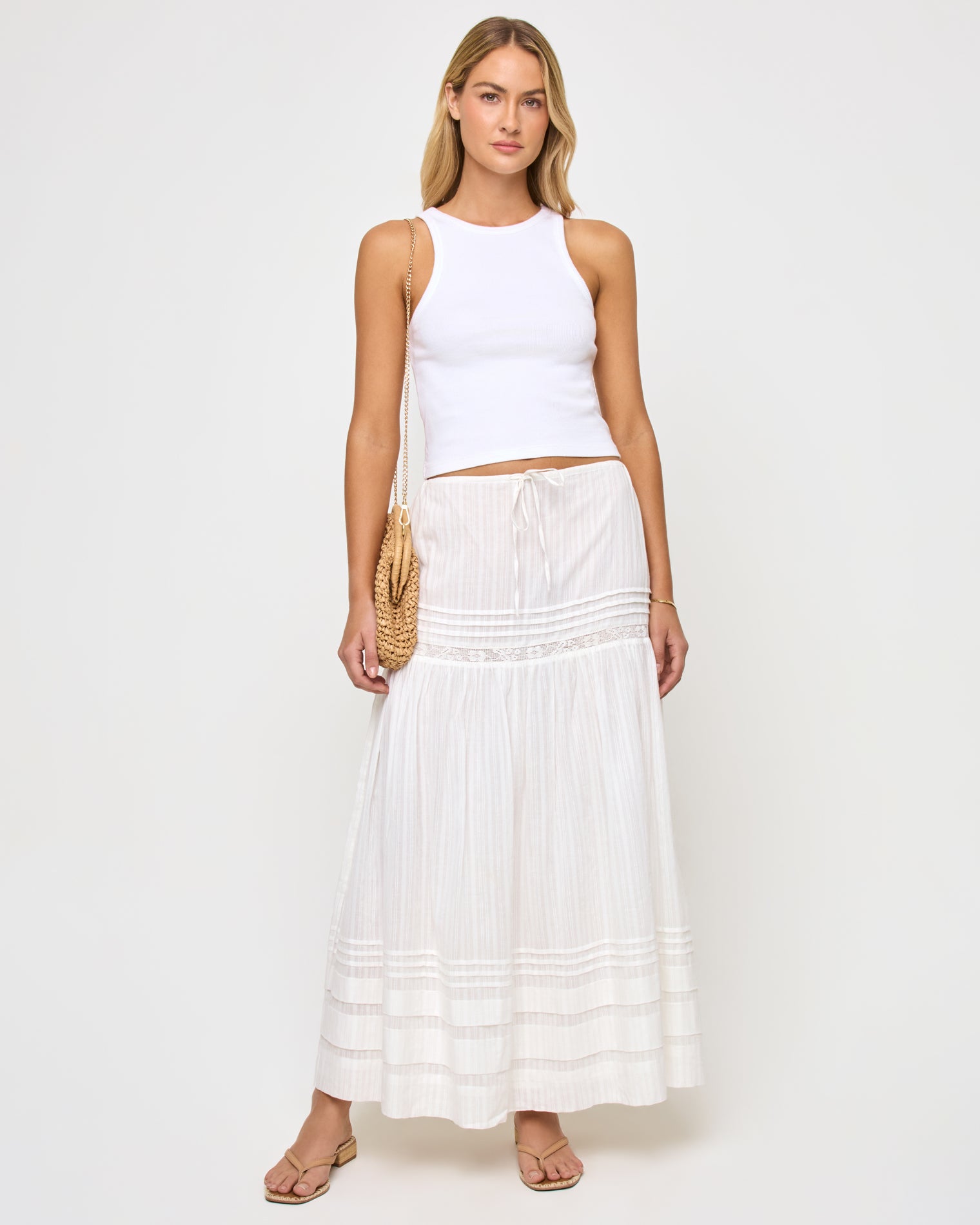 Reia Skirt - White Front view of a model wearing the LSPACE Reia Skirt in white, a tiered maxi skirt with pin-tuck and lace trim details, styled with a white crop top and straw shoulder bag, on a white studio background.