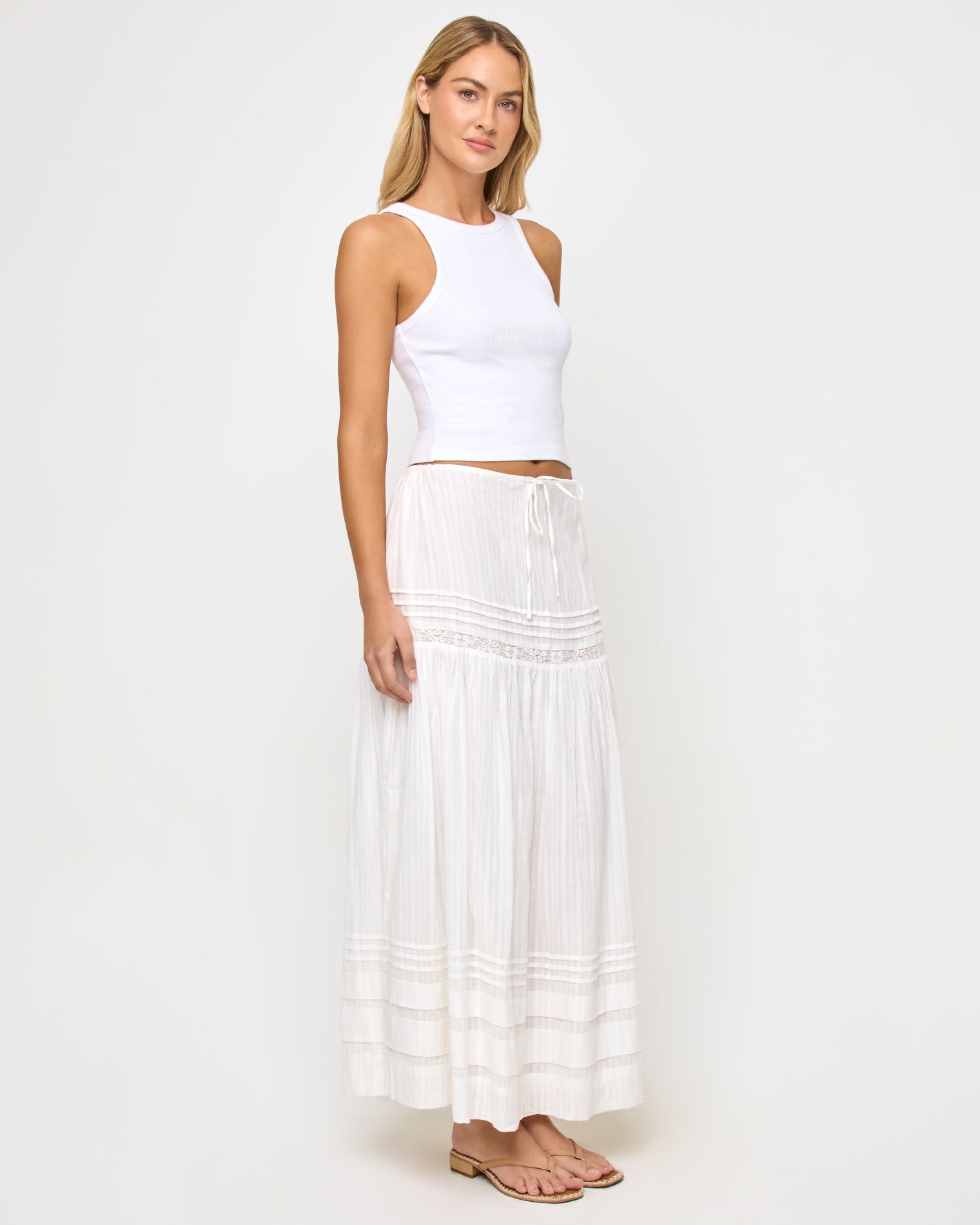 Reia Skirt - White Three-quarter front view of a model wearing the LSPACE Reia Skirt in white, a tiered maxi skirt with pin-tuck and lace trim details, styled with a white crop top, on a white studio background.