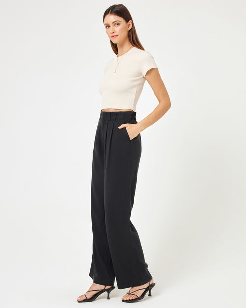 Side view of a model wearing the LSPACE Rhodes Pant in black, a high-waisted wide-leg trouser with front pleats, hand in pocket, styled with a white crop top and strappy heels.