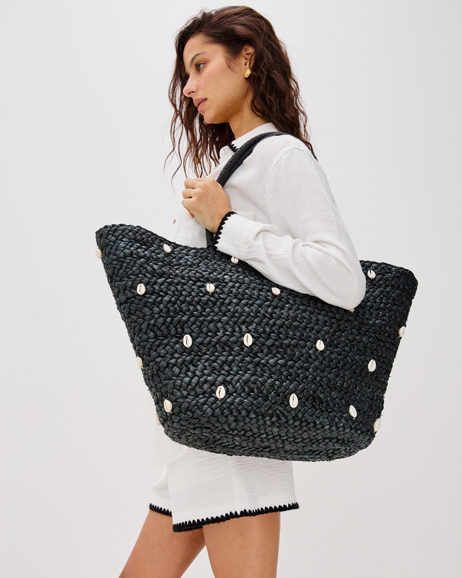 Baltic Seas Tote - Black Model carrying the Baltic Seas Tote in black over the shoulder, a large woven raffia tote bag with scattered cowrie shell embellishments, paired with a white button-down shirt and white skirt.